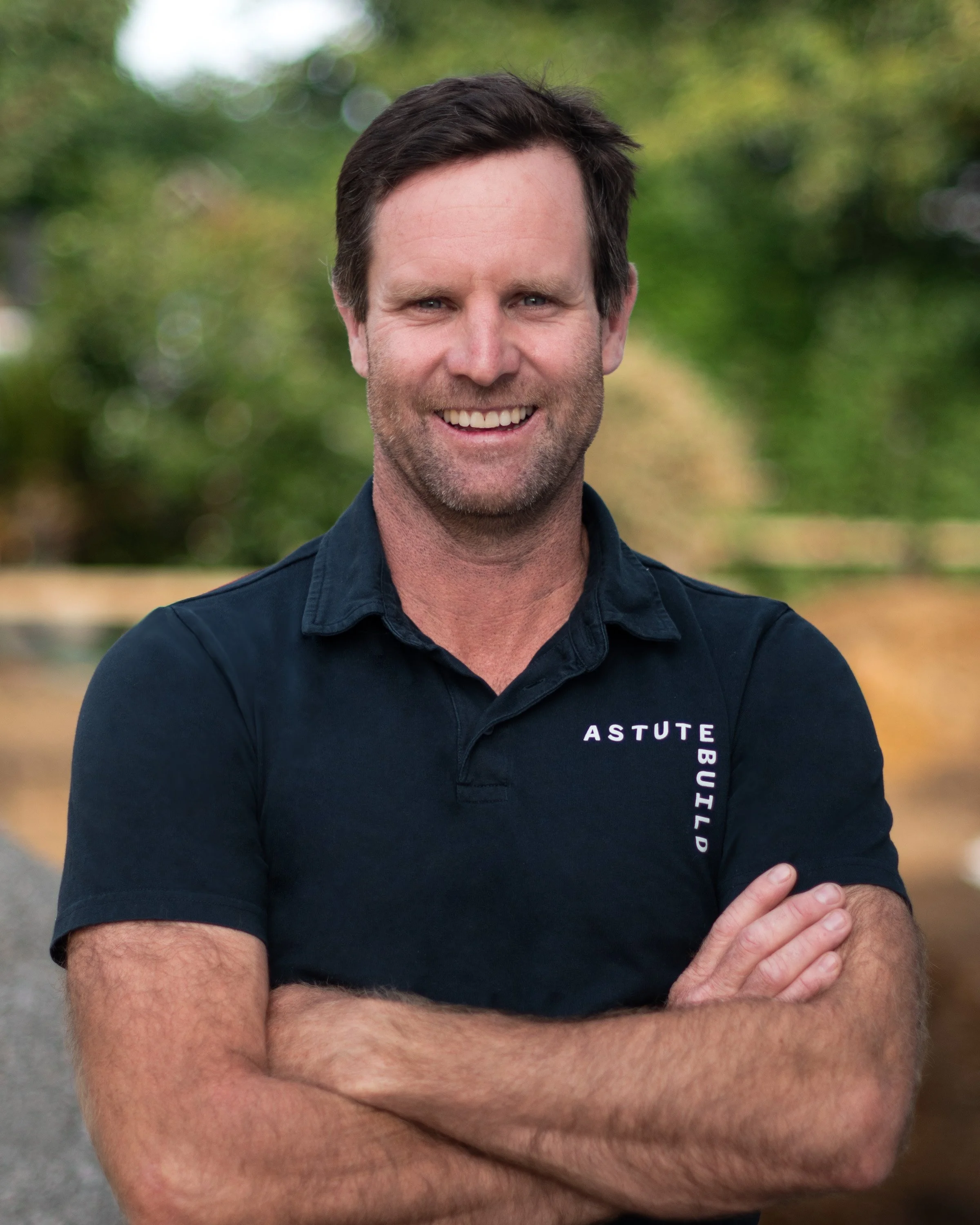 A smiling man with dark hair and a beard, wearing a dark blue polo shirt with the words 'ASTUTE BUILD' embroidered on it, standing outdoors with blurred trees and foliage in the background.