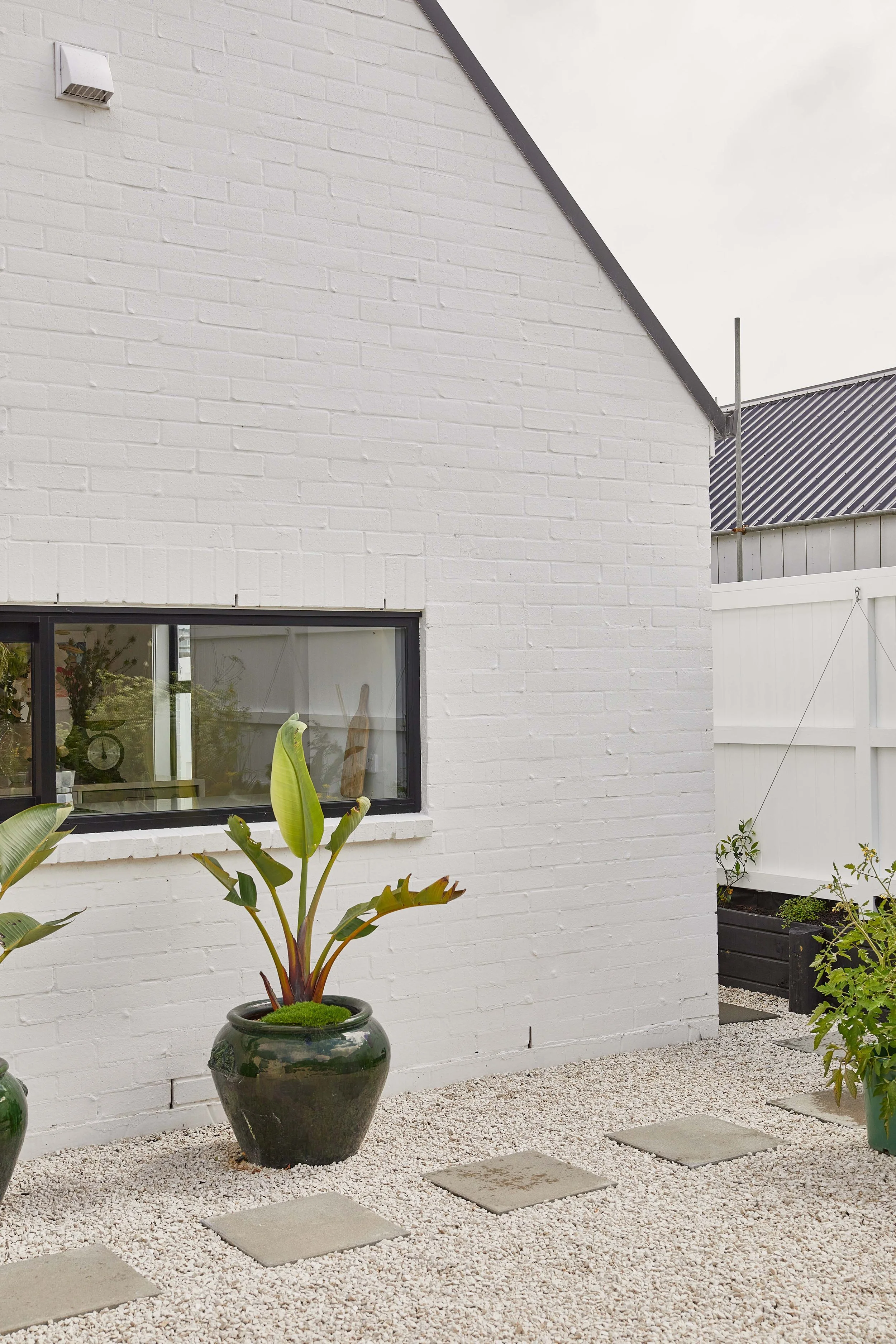 Exterior of a white brick house with a horizontal window, potted plants, and a gravel yard with stepping stones.