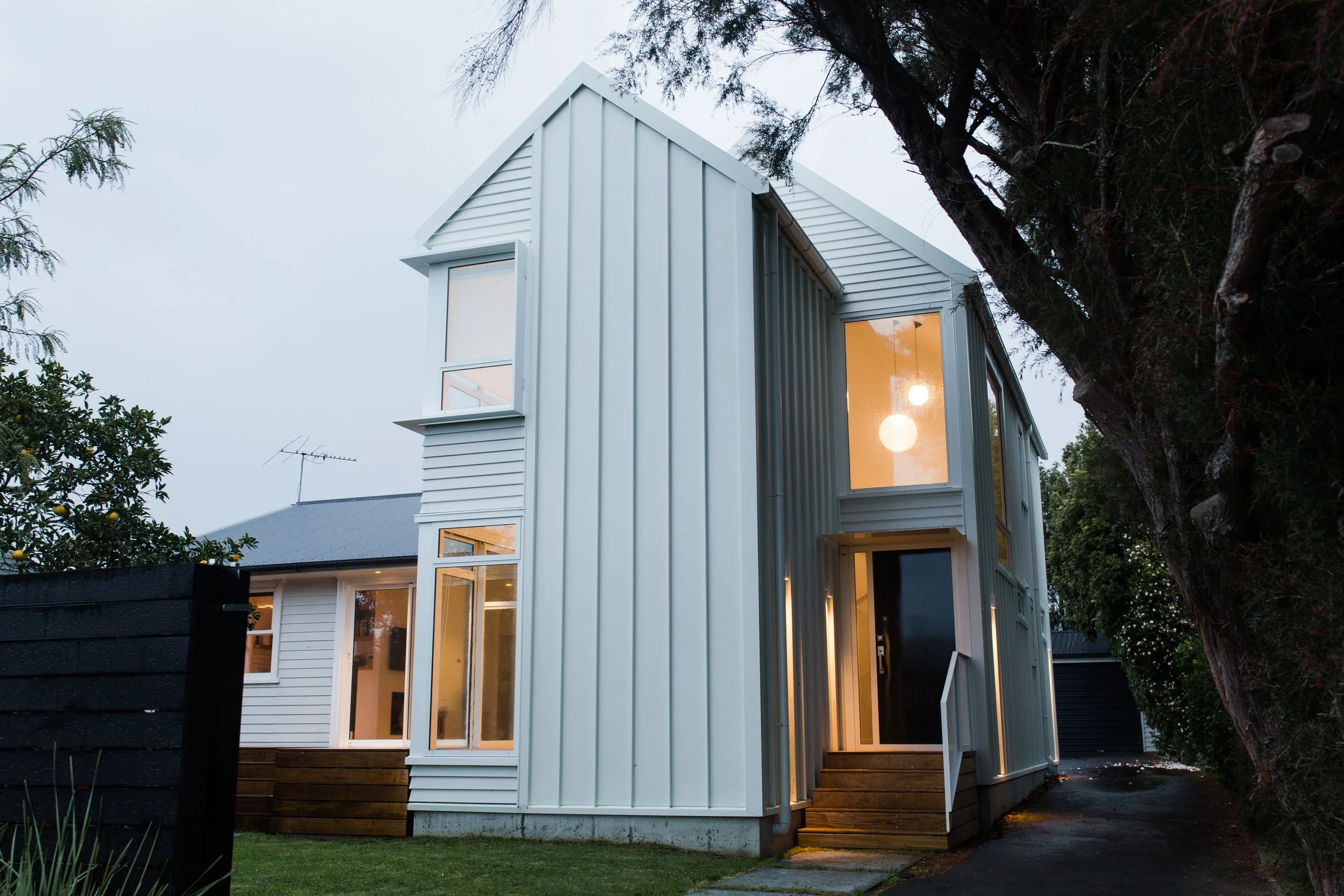 Modern two-story house with white siding and large windows, illuminated from inside, with trees and a narrow driveway adjacent, during dusk.