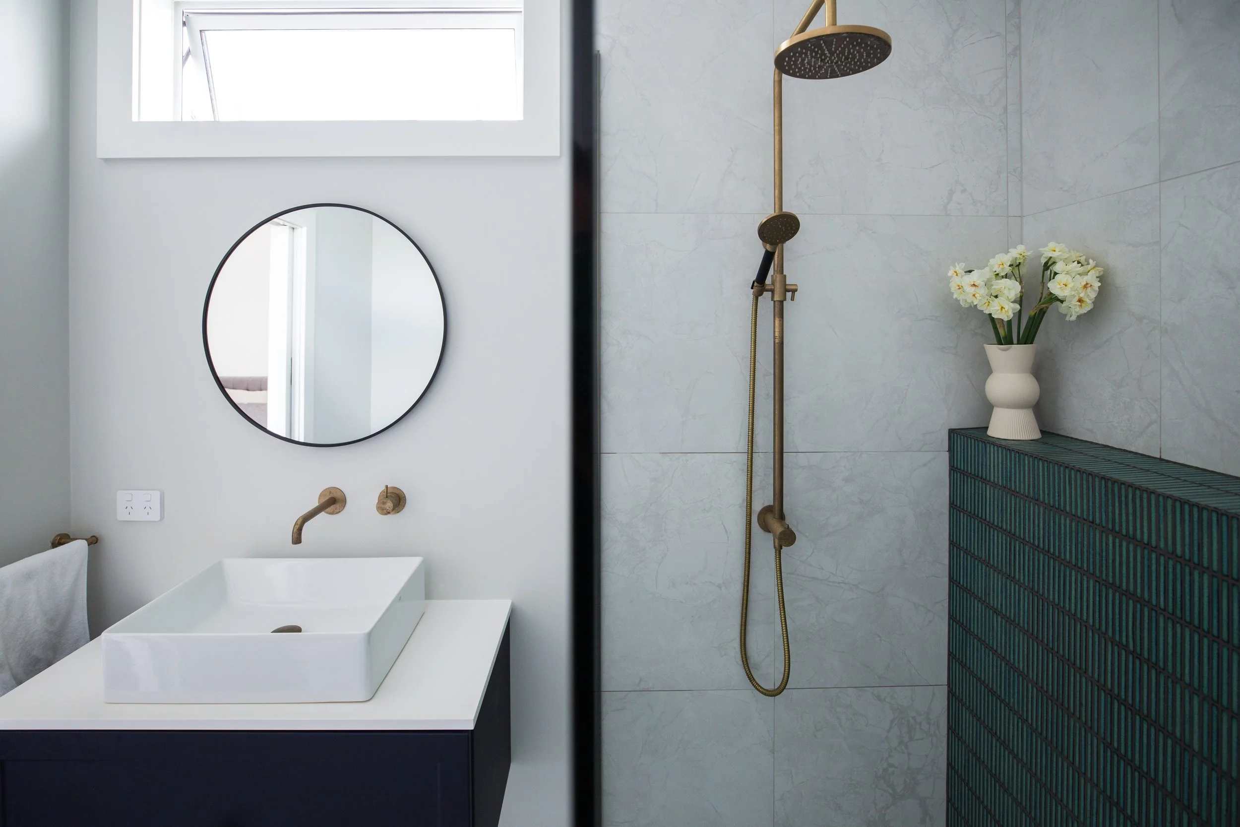 A bathroom with a white vessel sink, a round mirror, and a window above. The shower area features a brass showerhead and a vase with white flowers on a dark green tiled wall.
