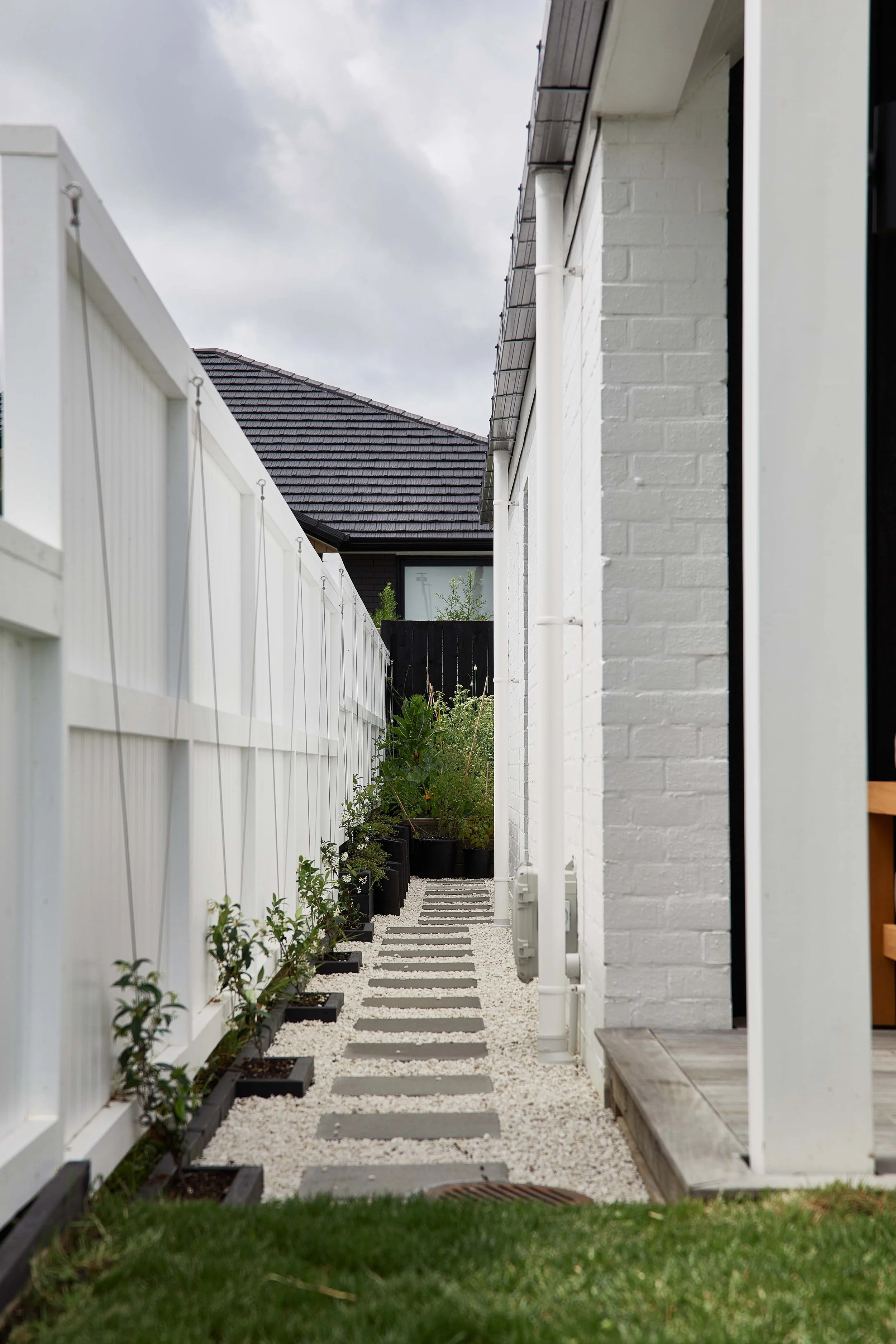 A narrow outdoor pathway bordered by a white fence on the left and a white brick house on the right, with potted plants and gravel on the path, and a cloudy sky overhead.