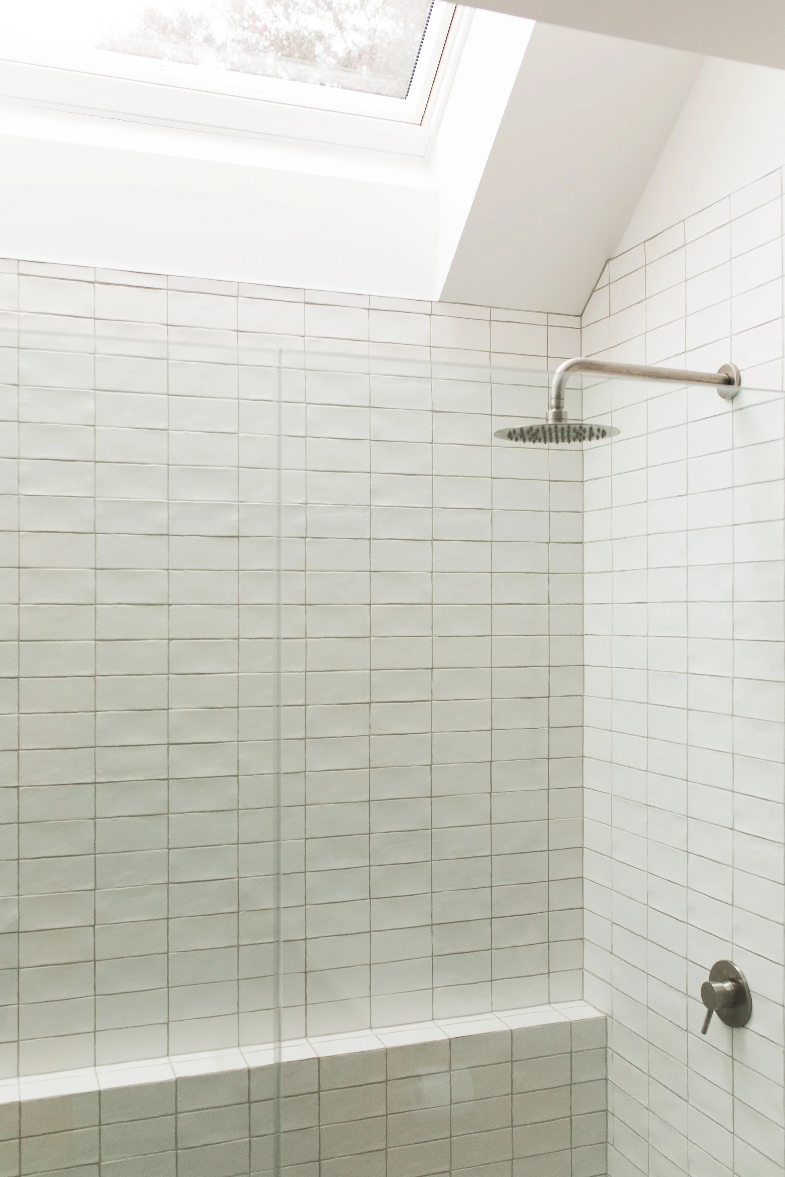 Modern shower with white tiled walls, a rain showerhead, a glass shower door, and a skylight window.