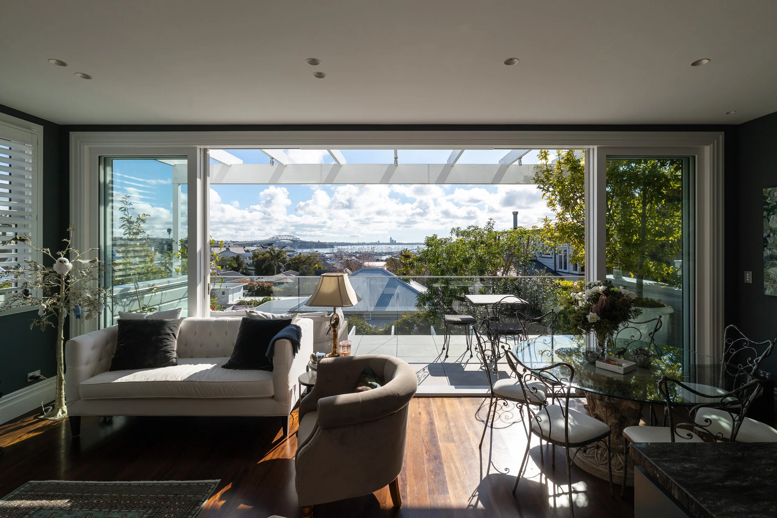 Bright living room with large sliding glass doors opening to a balcony view of a cityscape, blue sky, trees, and rooftops.