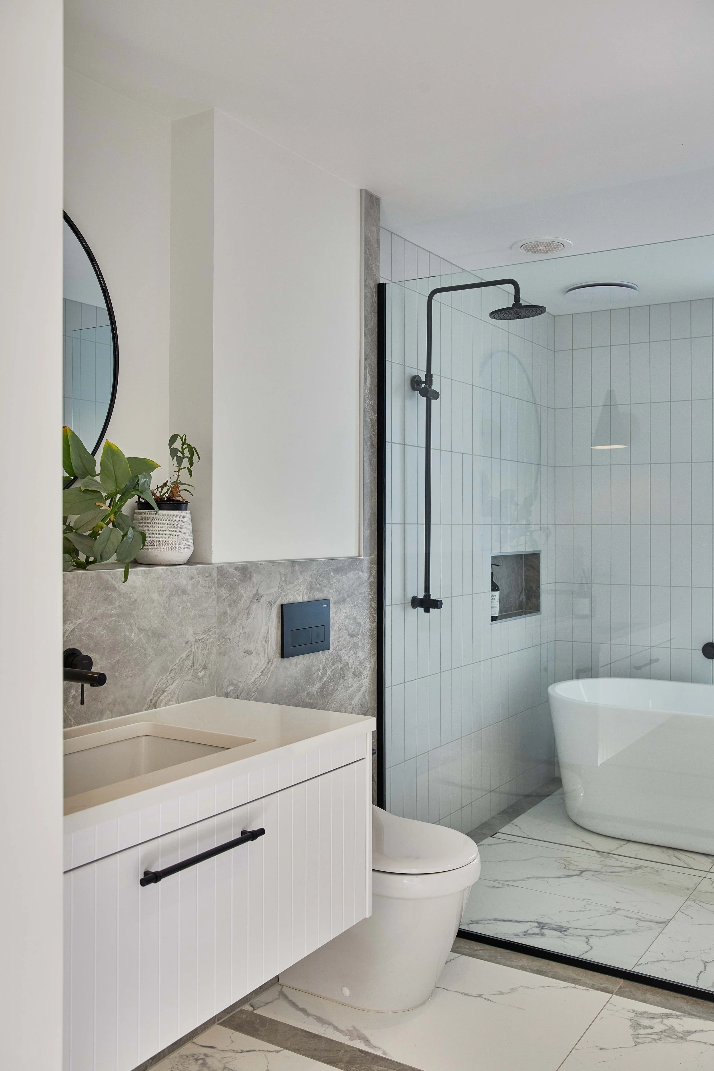 Modern bathroom with white vanity, wall-mounted toilet, shower with black fixtures, and a bathtub, decorated with potted plant, mirror, and marble flooring.