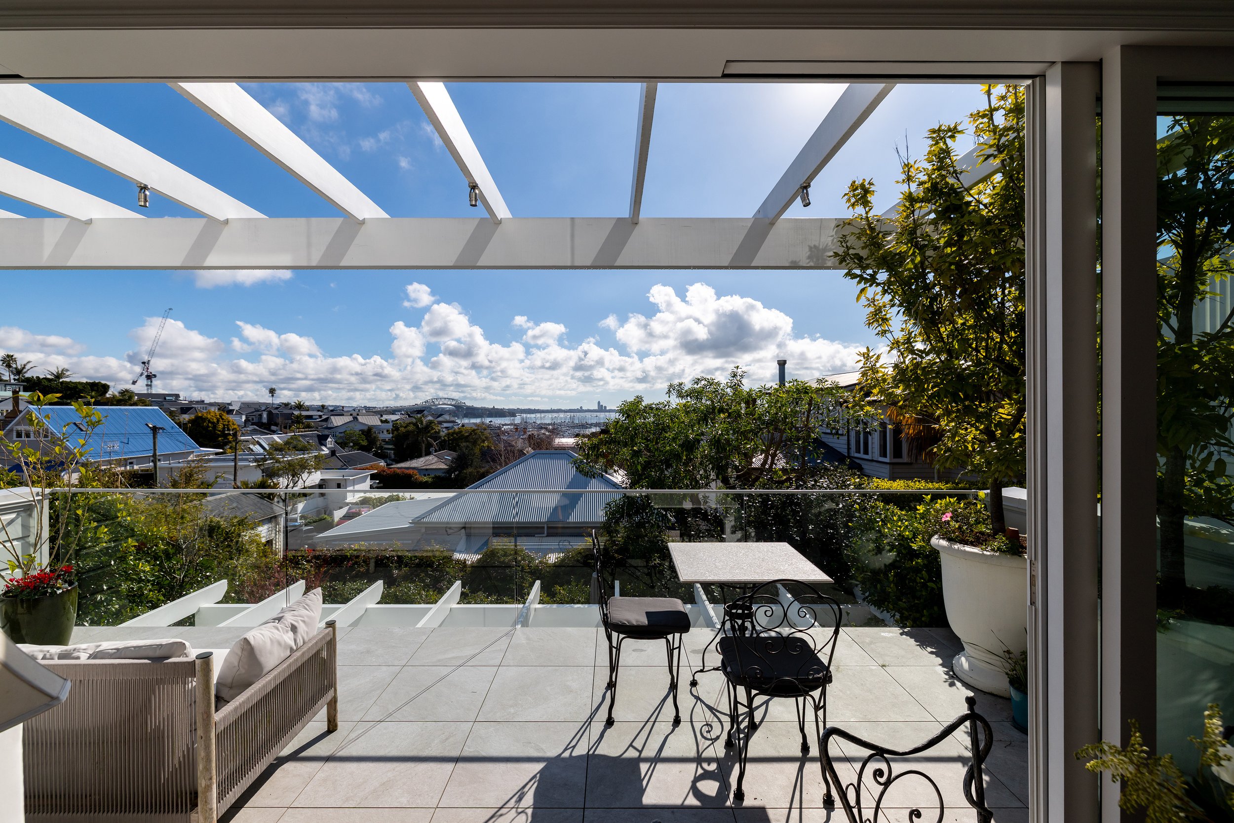 View of a rooftop balcony with outdoor furniture, potted plants, trees, and a cityscape with clouds and blue sky in the background.