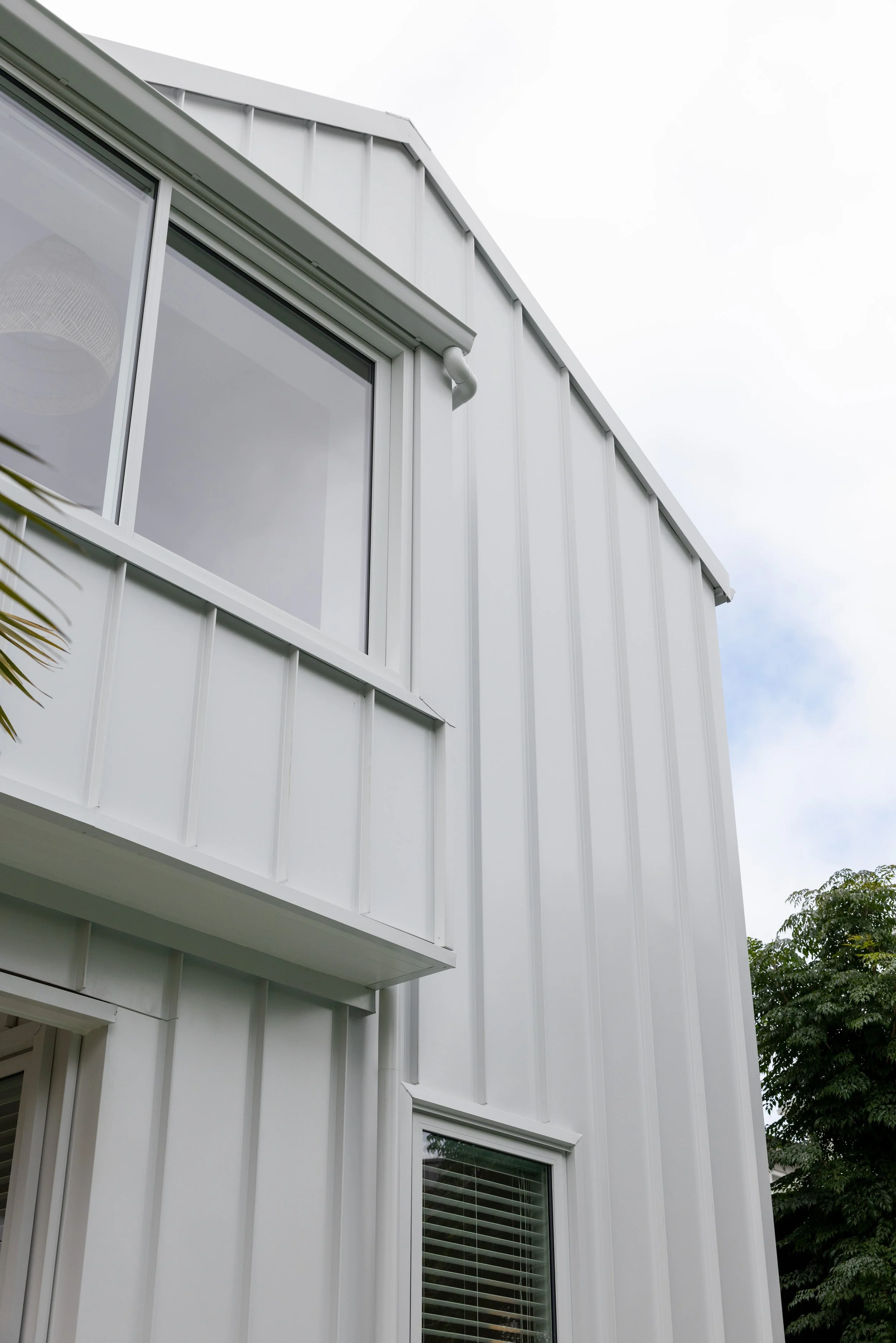 Close-up of a modern white metal building with vertical siding, large windows with blinds, and a portion of a balcony, with a cloudy sky and green trees in the background.