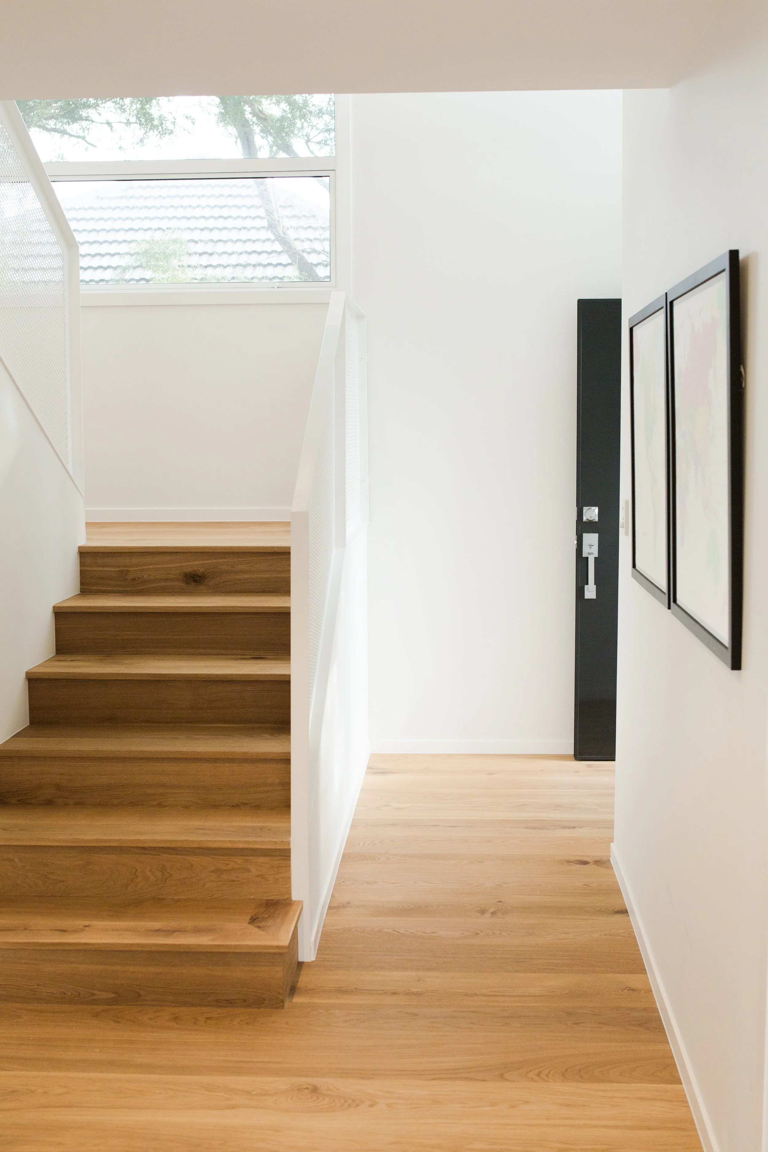 Interior view of a modern home with wooden stairs, hardwood flooring, and a large window letting in natural light, with framed artwork on the wall.