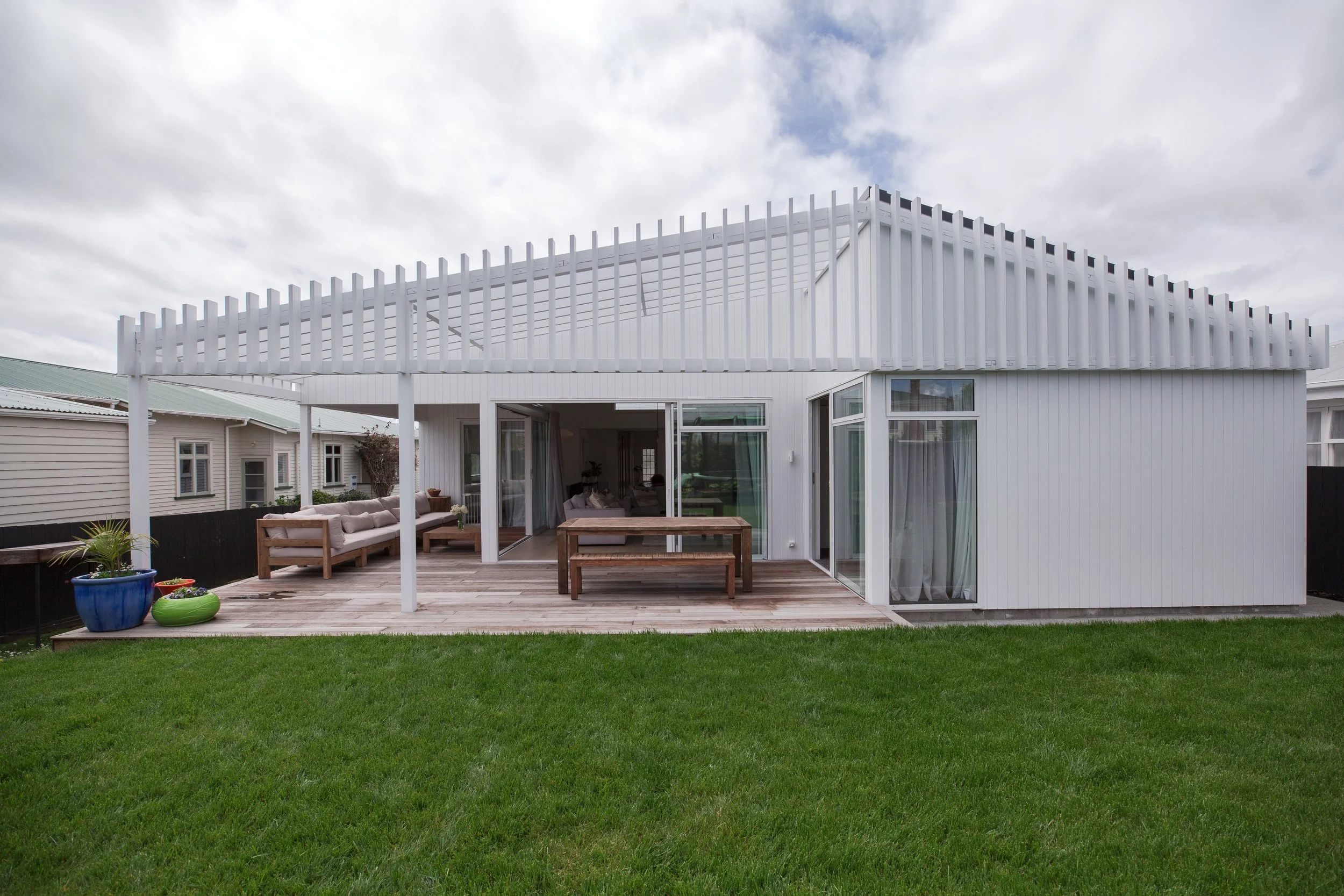 White modern house with a wooden deck, outdoor seating, and potted plants, surrounded by a green lawn under a cloudy sky.