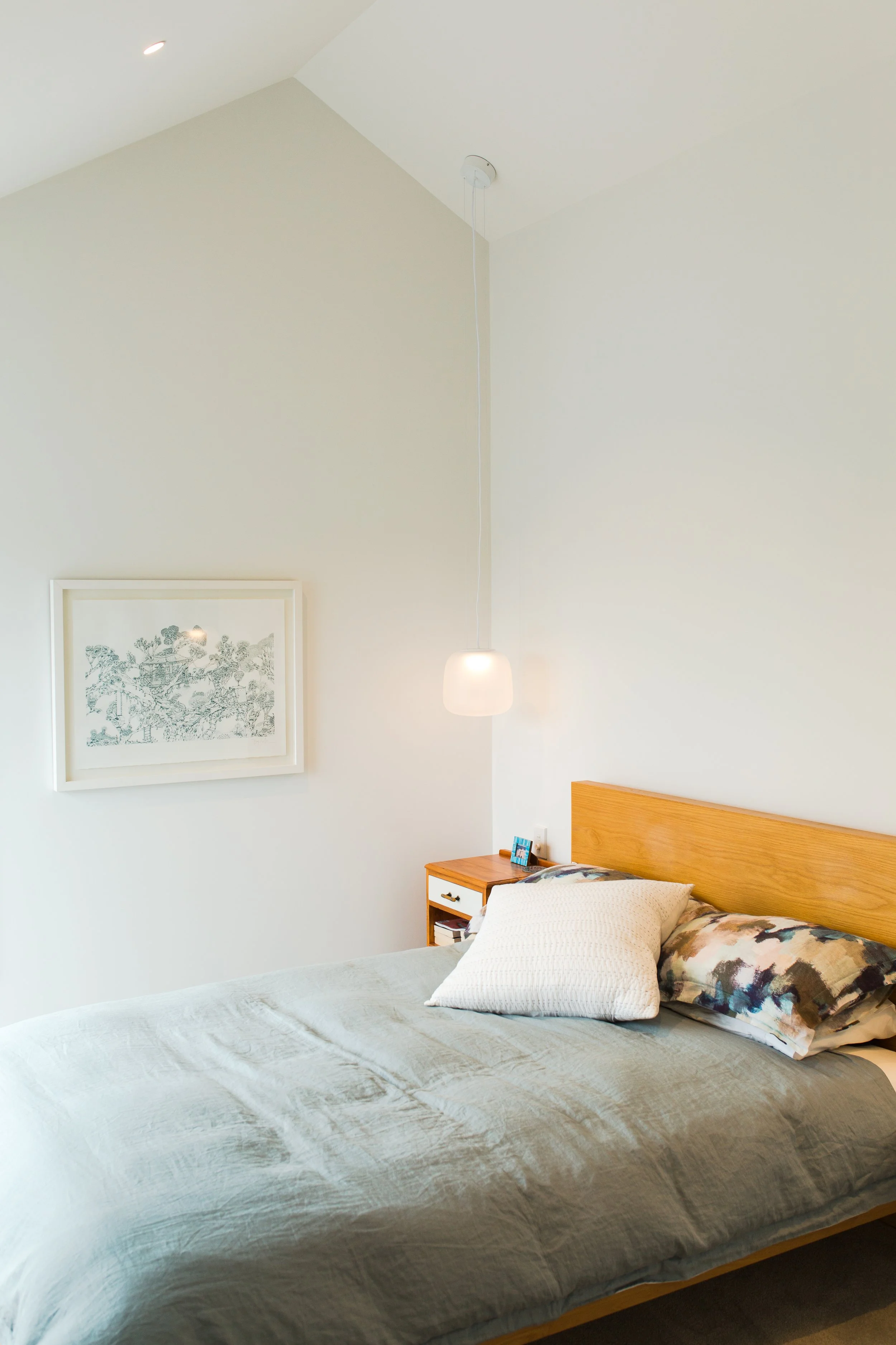 A bedroom with a bed, wooden headboard, pillows, a nightstand with a small framed photo, a hanging light, and a framed artwork on a white wall.