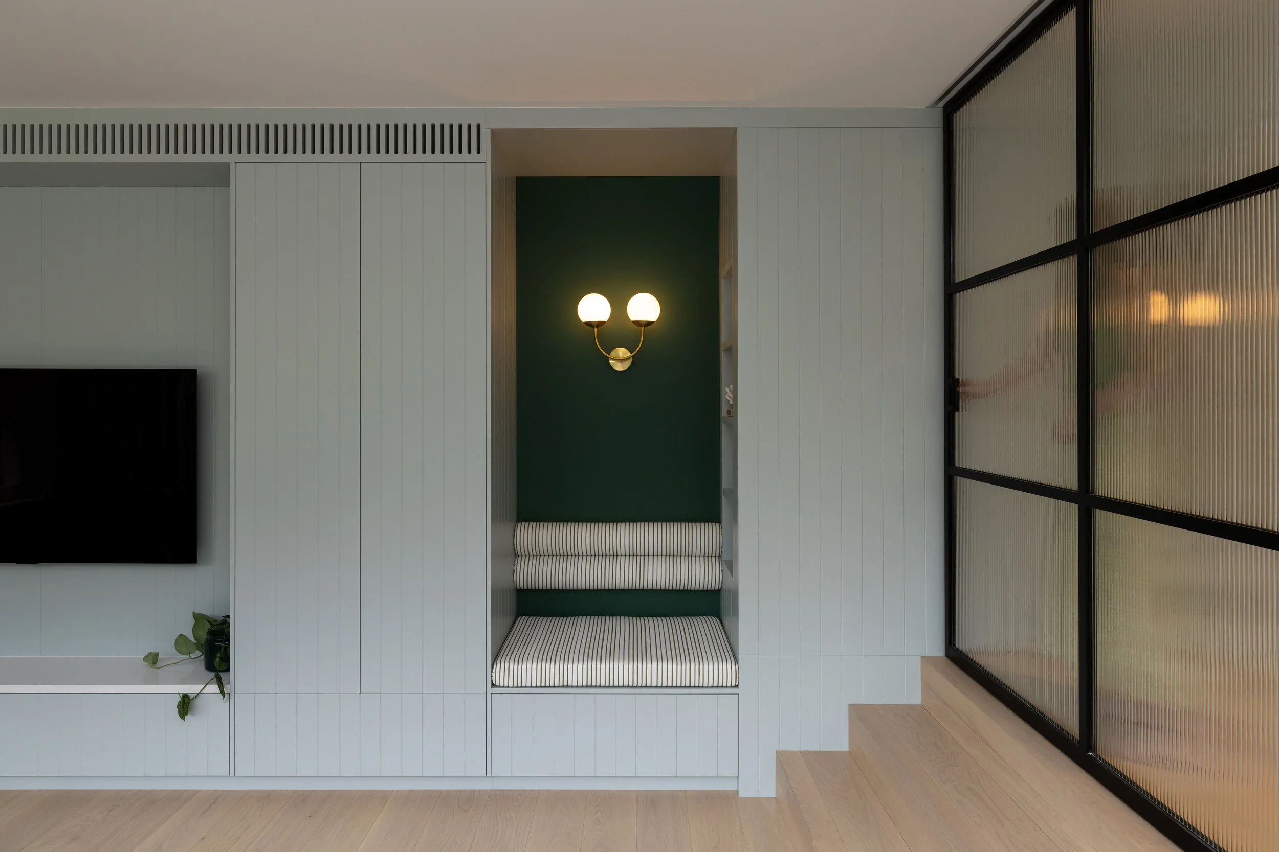 Interior view of a modern living room with a wall-mounted TV, built-in seating nook with striped cushions, green accent wall, wall-mounted light fixture, and frosted glass sliding door.