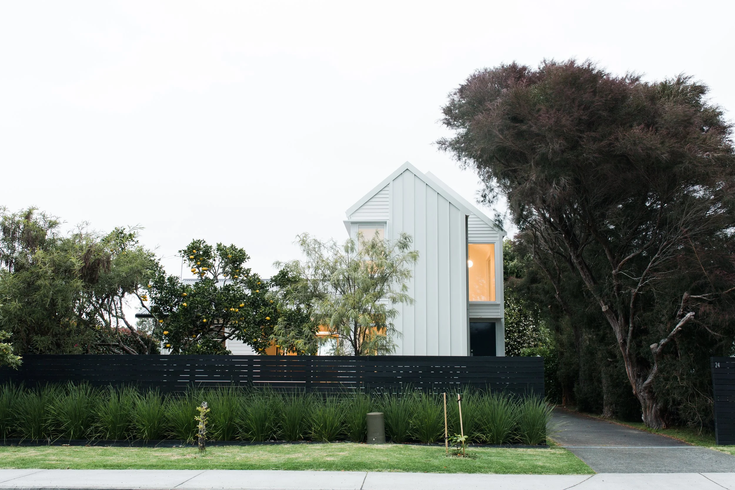 Modern white house with a steep roof, large windows, surrounded by trees and green plants, with a black fence in front.