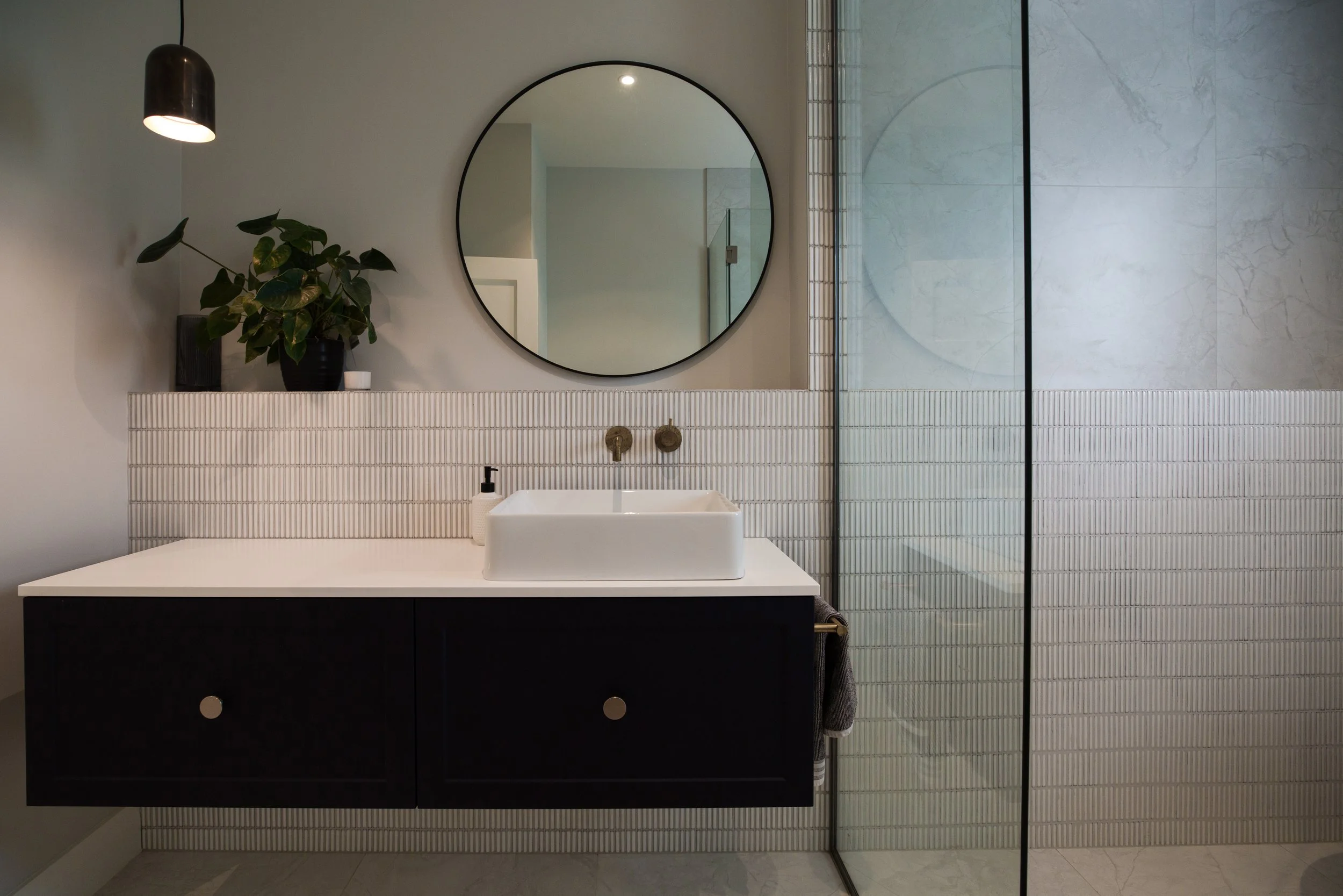 Modern bathroom with a white countertop, black cabinet, round mirror, white vessel sink, black and gold fixtures, potted plant, and glass-enclosed shower with marble walls.