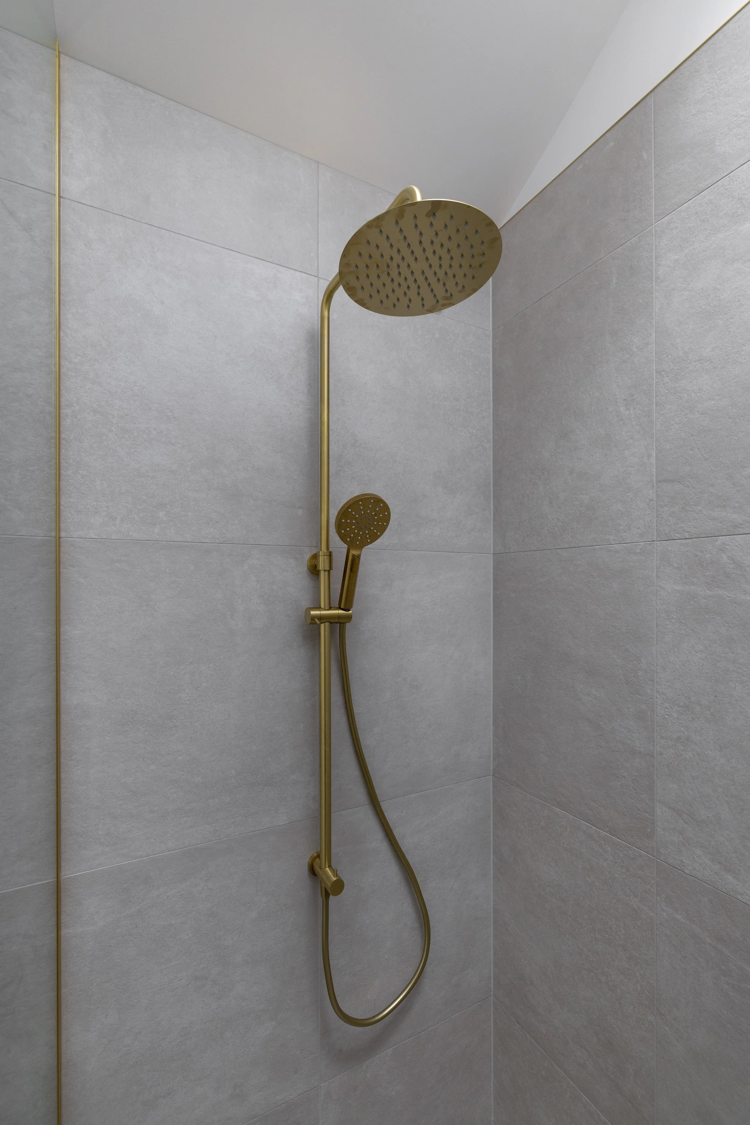 A modern shower with a brass showerhead and a handheld shower wand mounted on the wall in a tiled shower stall.