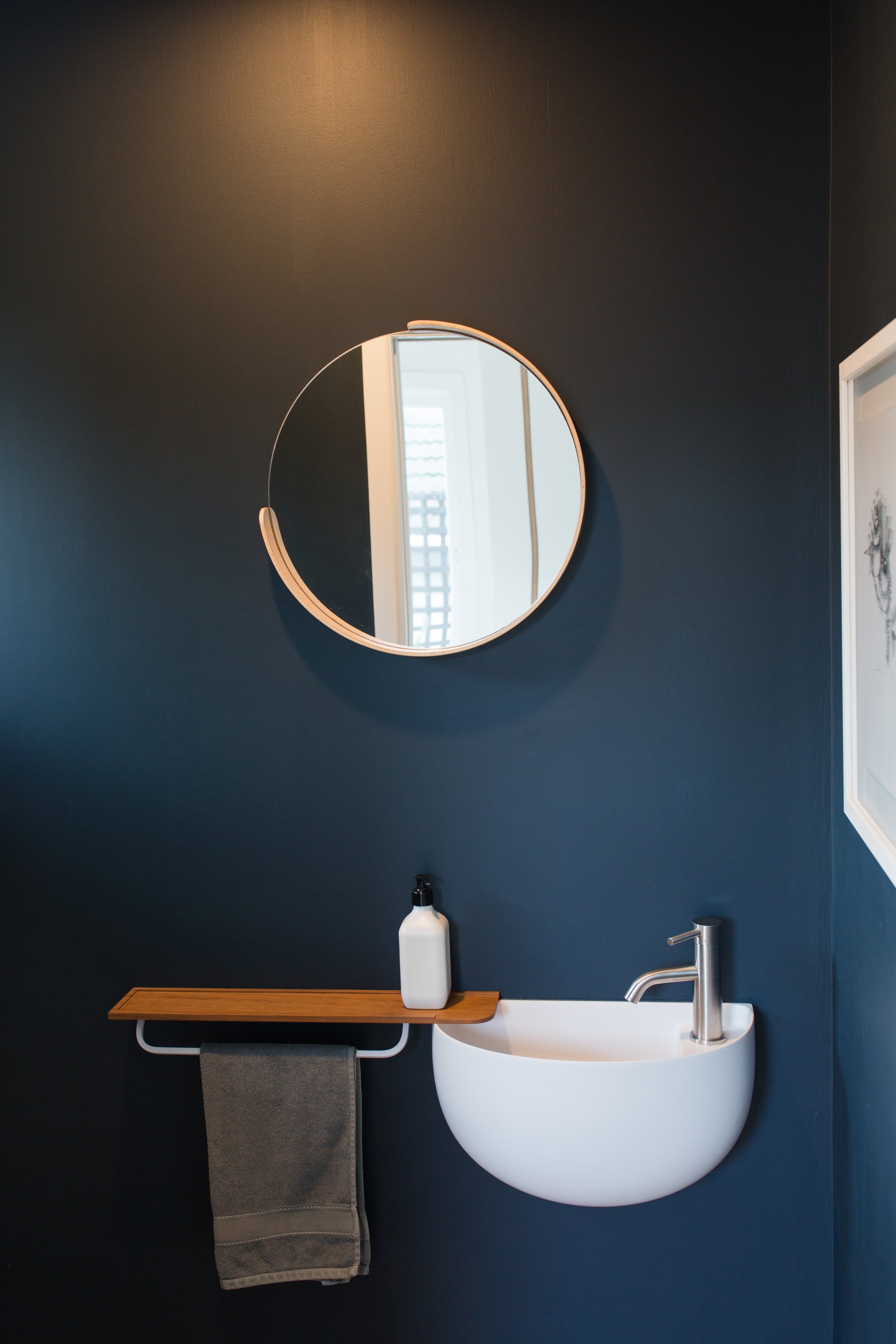 Modern bathroom with navy blue wall, round mirror, small white sink, soap dispenser, and a wooden towel rack with a gray towel.