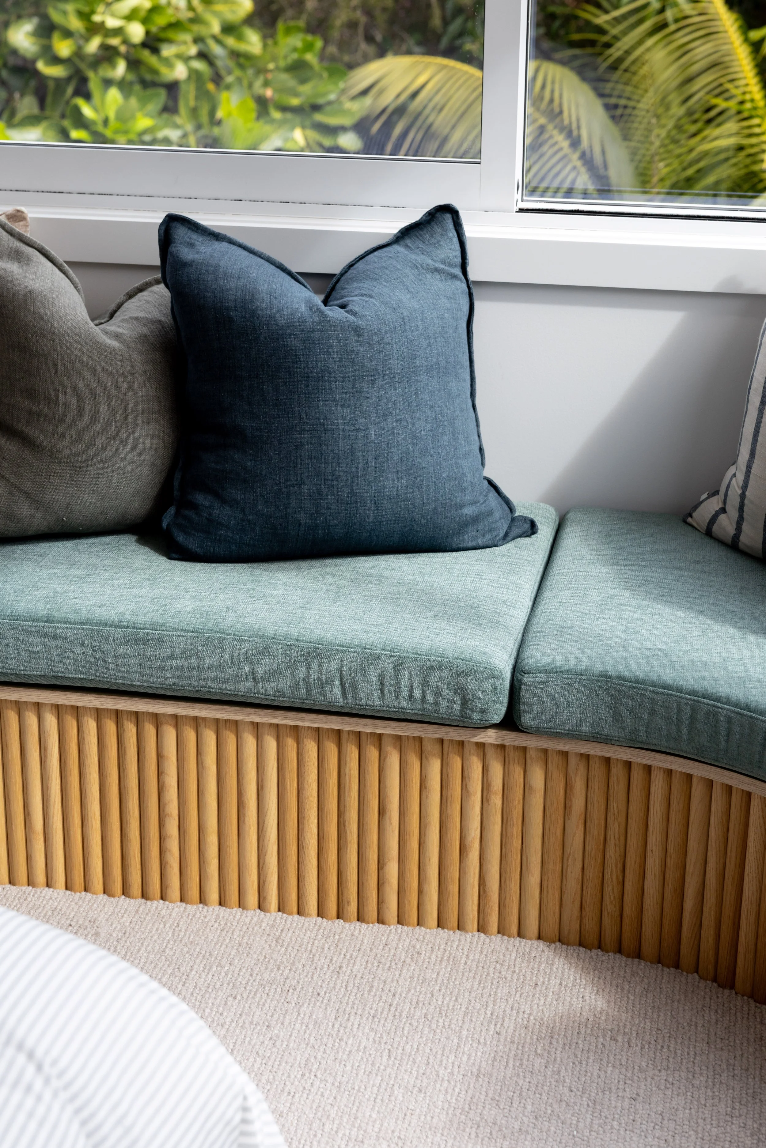Close-up of a window seat with light blue cushions and decorative pillows in front of a window with greenery outside.