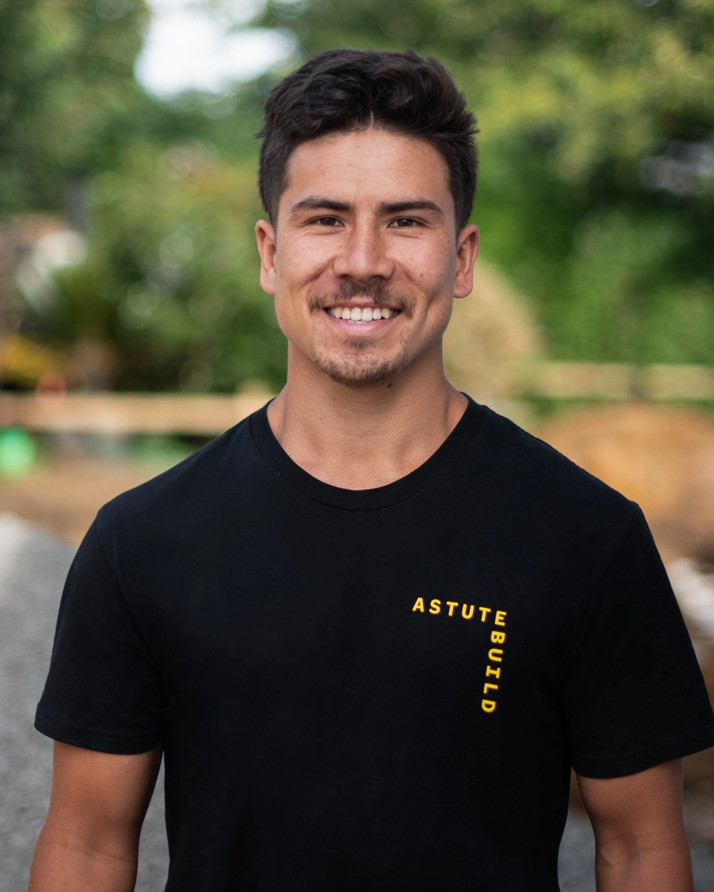 A young man with dark hair, smiling, wearing a black T-shirt with yellow text on the chest that says 'ASTUTE BUILD', outdoors with green trees and a blurred background.