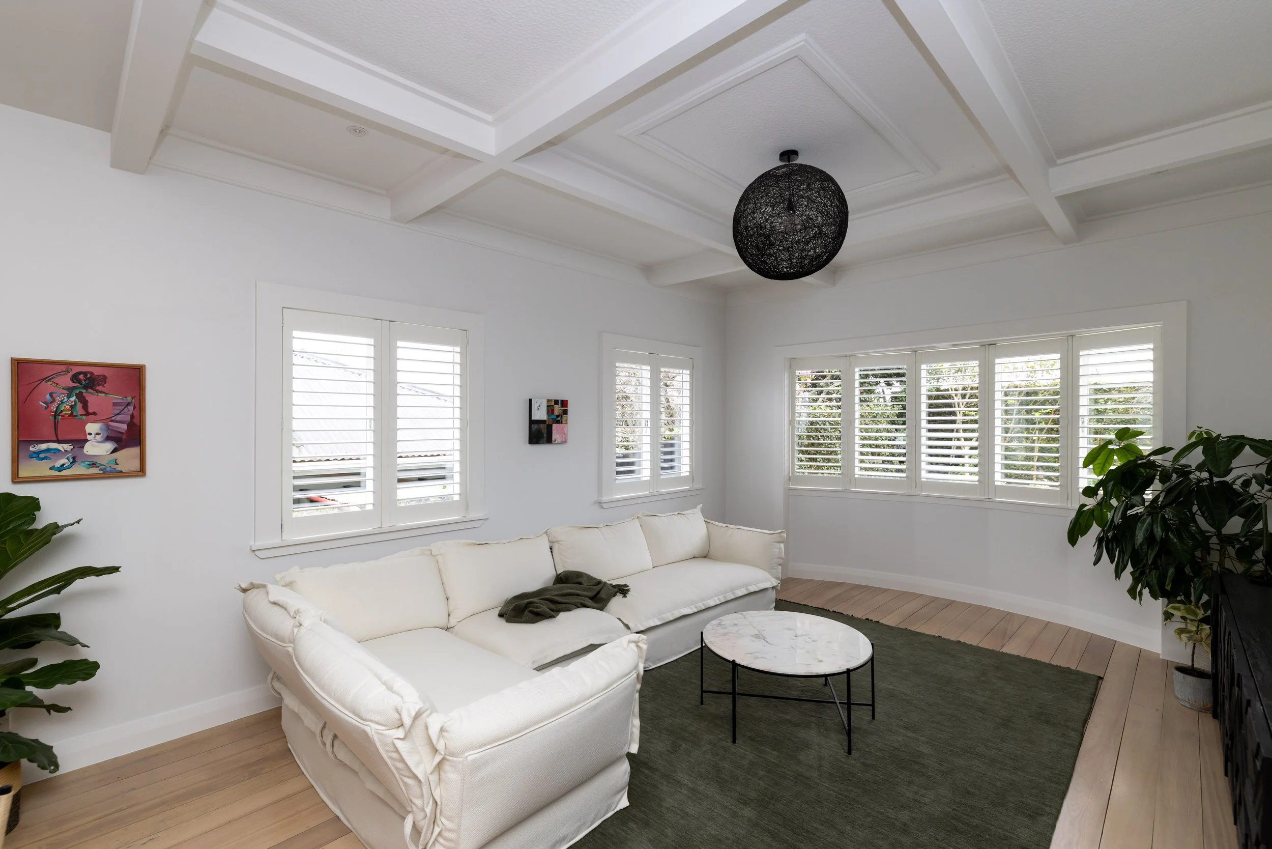 Living room with white sectional sofa, round marble coffee table, green rug, window shutters, plants, and artwork on the white walls.