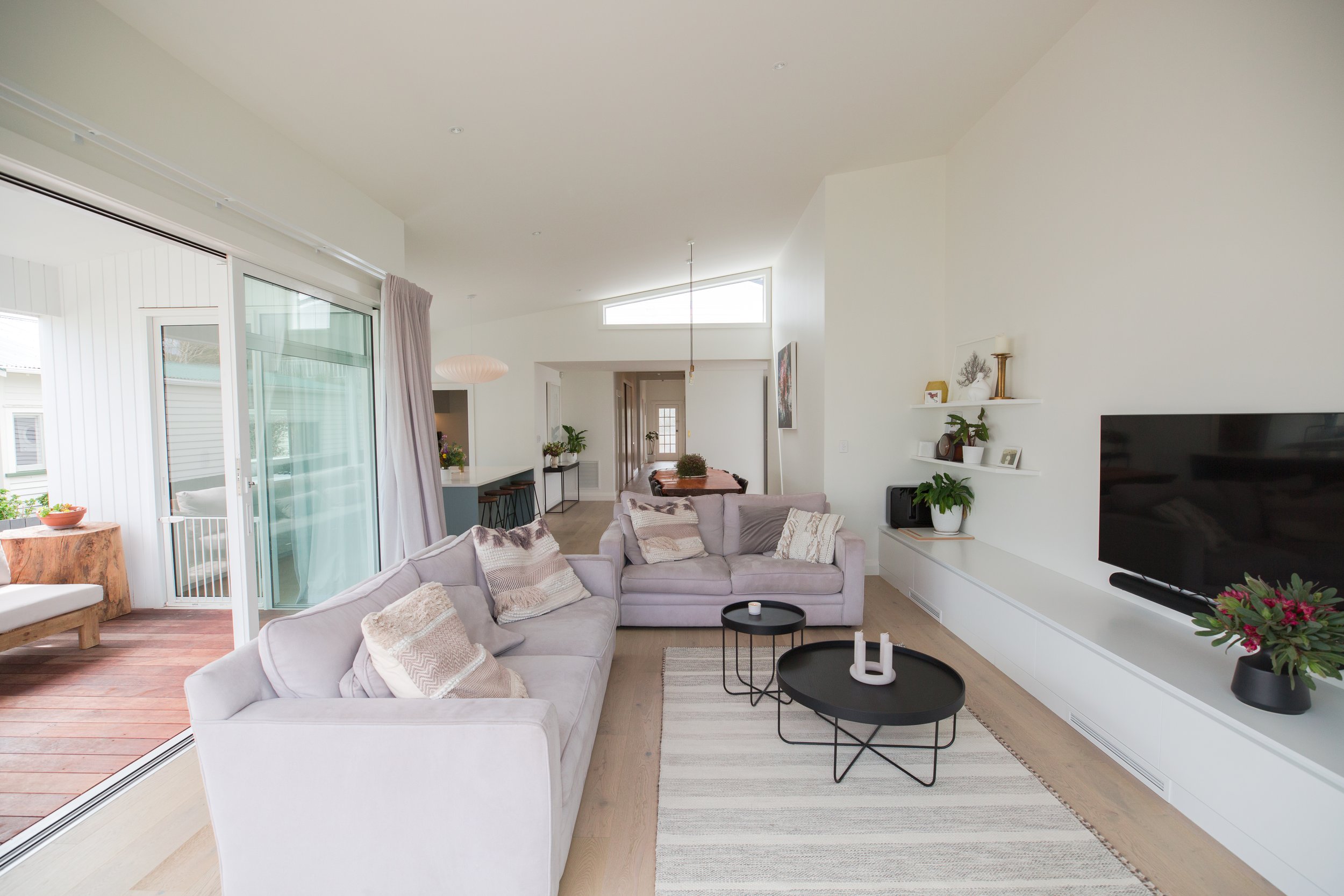 Bright living room with two light-colored sofas, black coffee tables, a rug, a TV on the wall, and a sliding glass door leading to an outdoor patio.