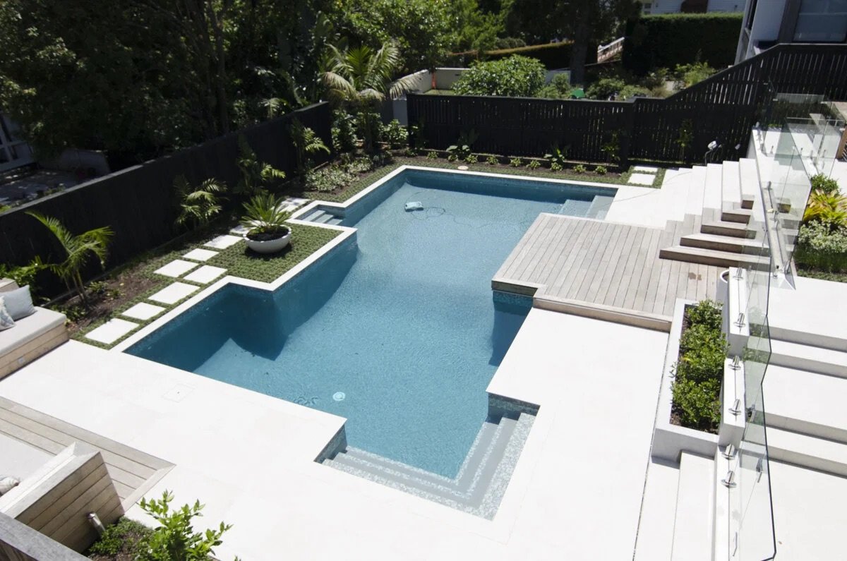 A modern backyard swimming pool with a shallow, elevated spa area and a connected deep swimming section, surrounded by a wooden deck, potted plants, and a garden with shrubs and trees.