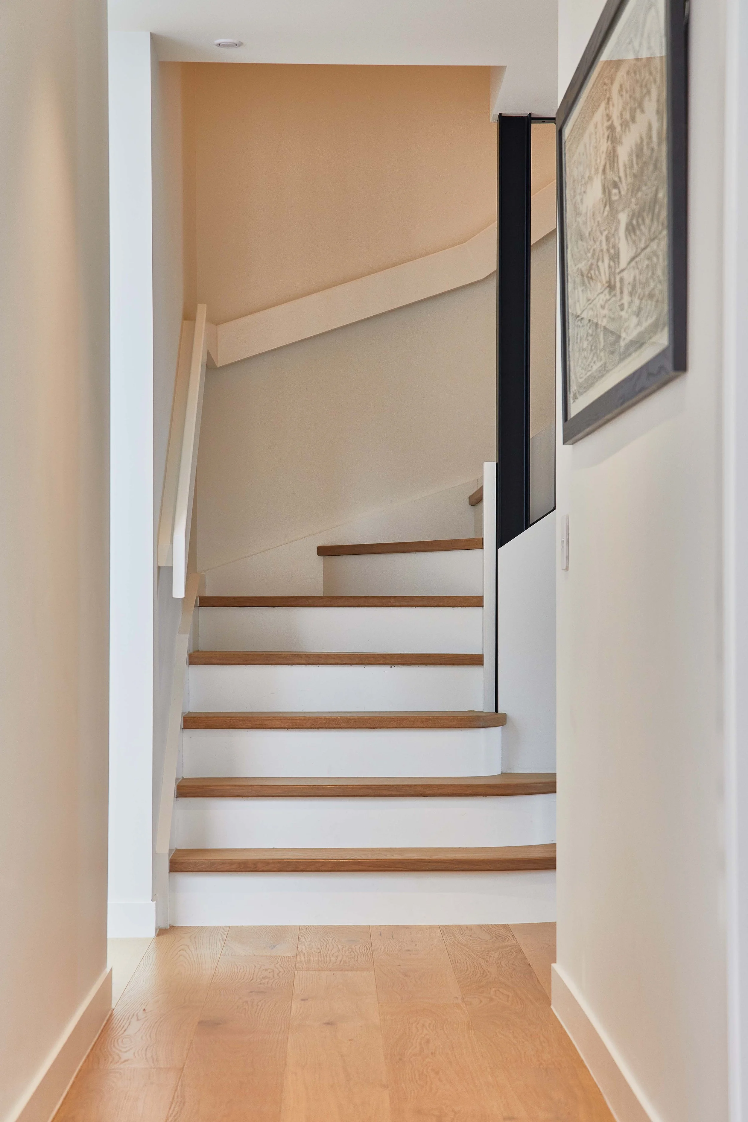 View of a staircase with wooden steps and white risers, seen from a hallway with a wooden floor. A framed artwork is partially visible on the right wall, and a black and white wall feature is on the right side of the staircase.