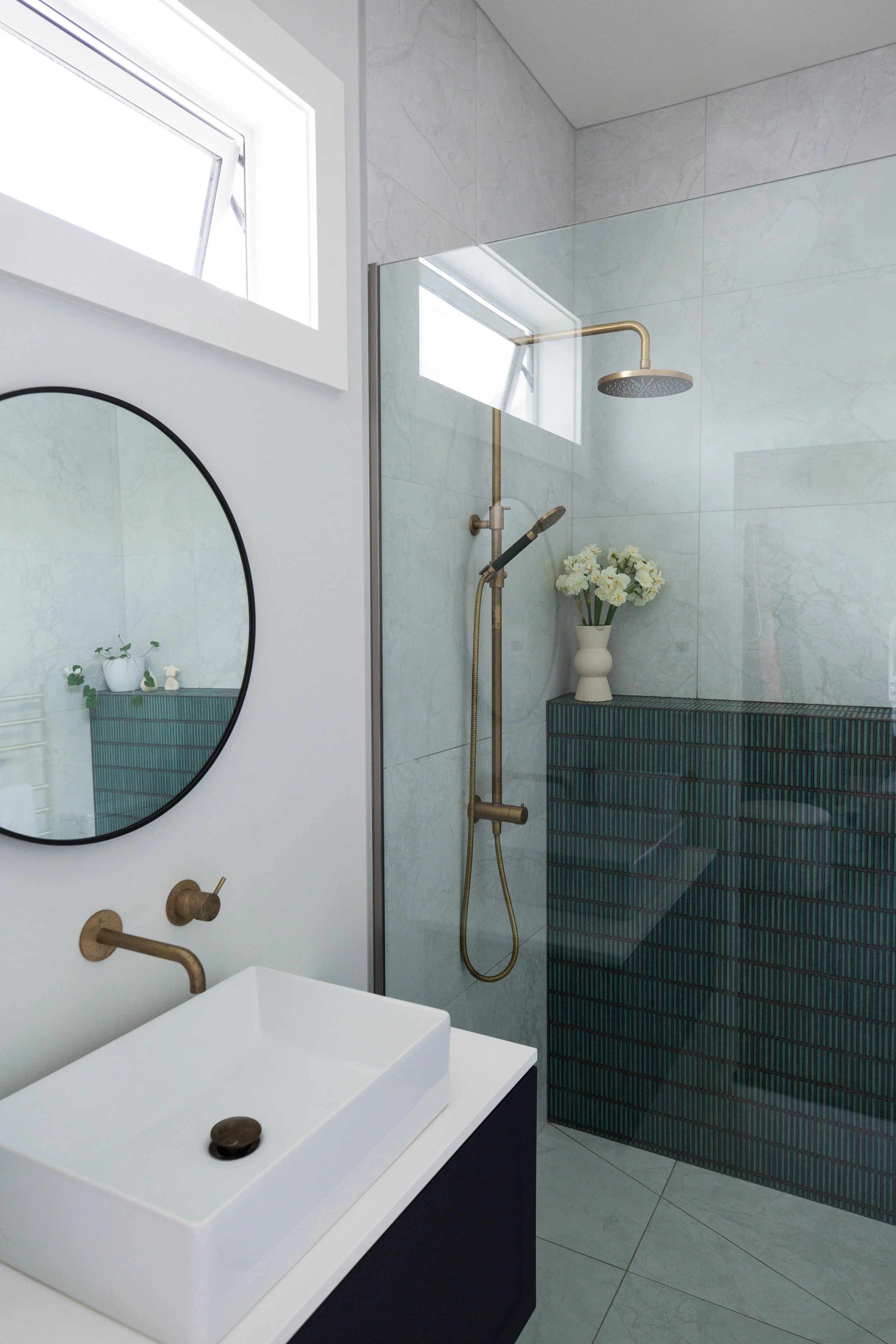 Modern bathroom with a vessel sink, round mirror, and a walk-in shower with brass fixtures and a flower vase, window with natural light.