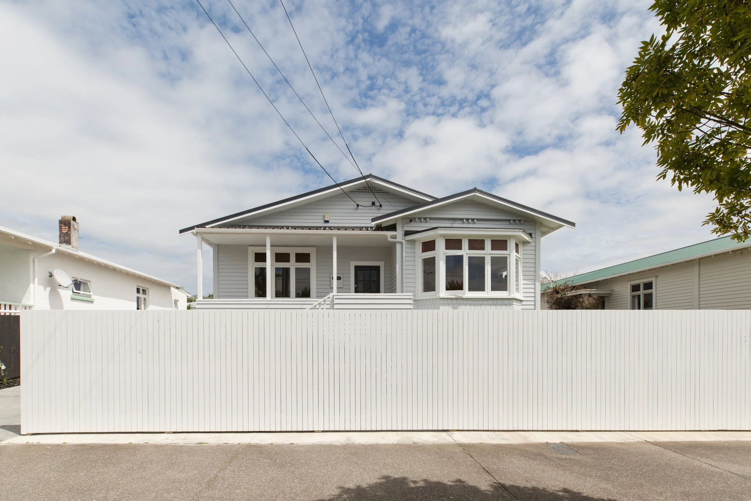 Front view of a white house with a raised porch, bay window, and a vinyl fence across the front. The sky is partly cloudy with some blue visible.
