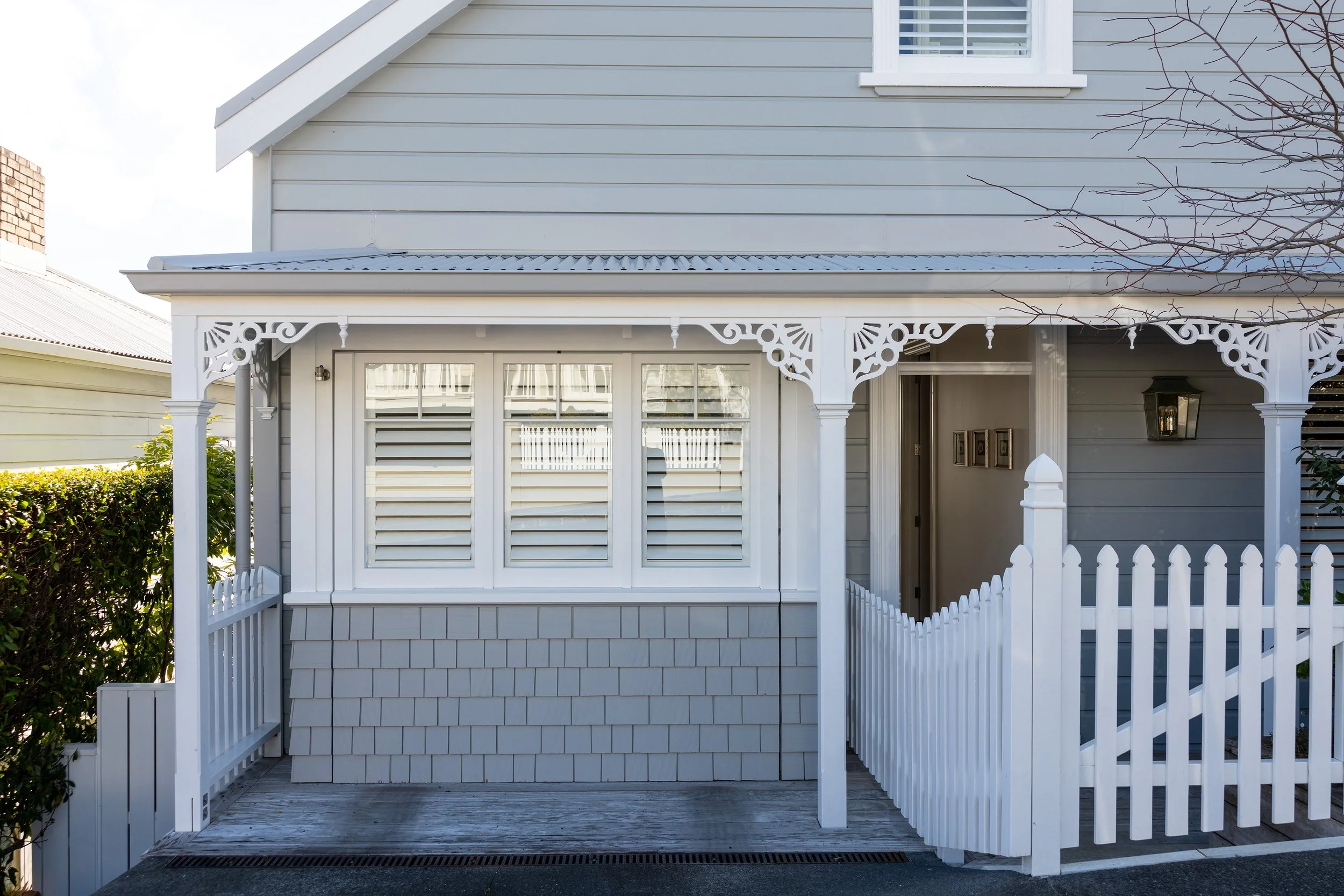 Front view of a light gray house with white decorative trim, windows with white shutters, a small porch with a white picket fence, and a large lantern-style light fixture next to the entrance door.