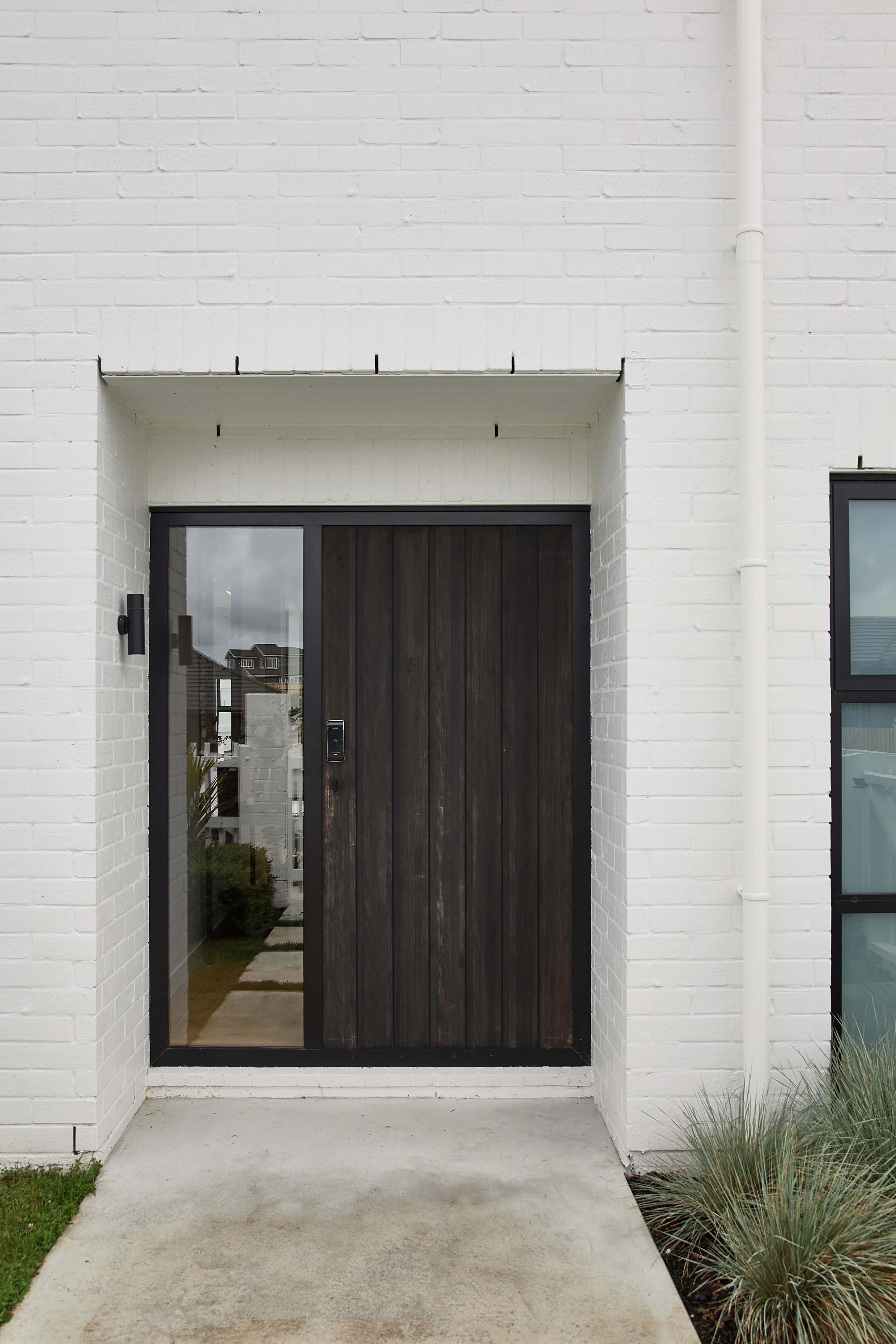 Modern house entrance with a dark wooden front door on a white brick building, glass sidelights, and a small concrete porch.