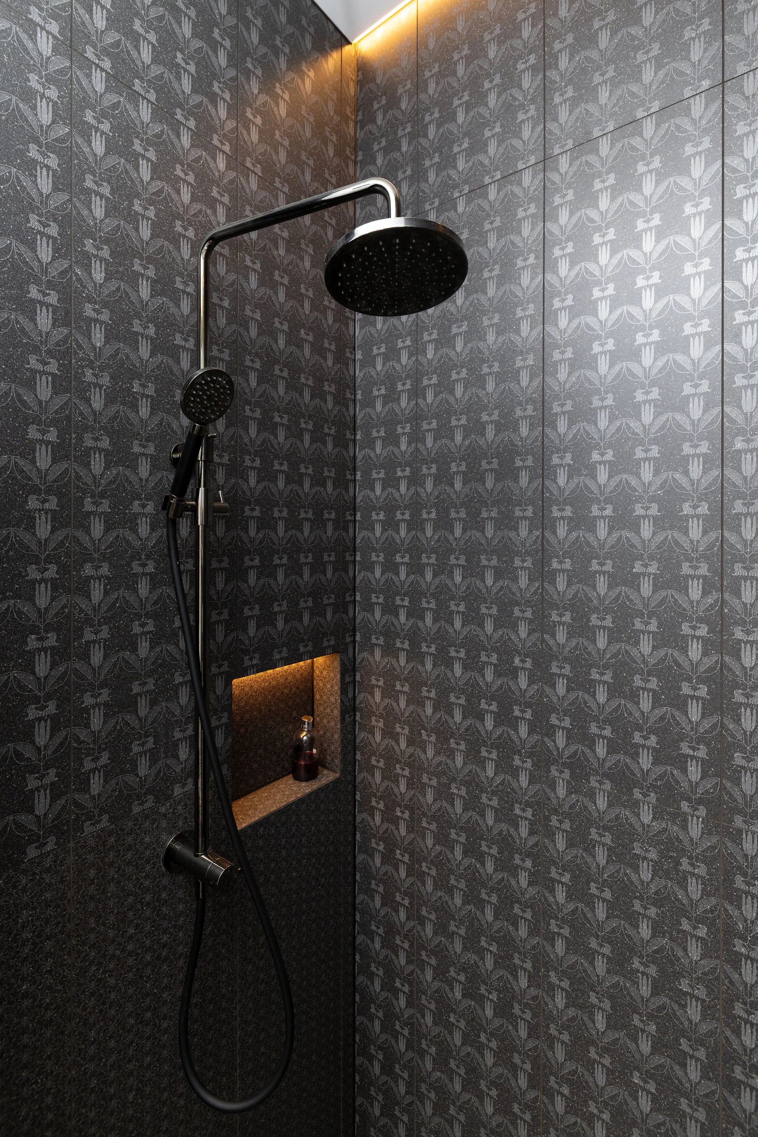 Dark tiled shower with black showerheads, built-in shelf with a bottle, and ceiling lights.