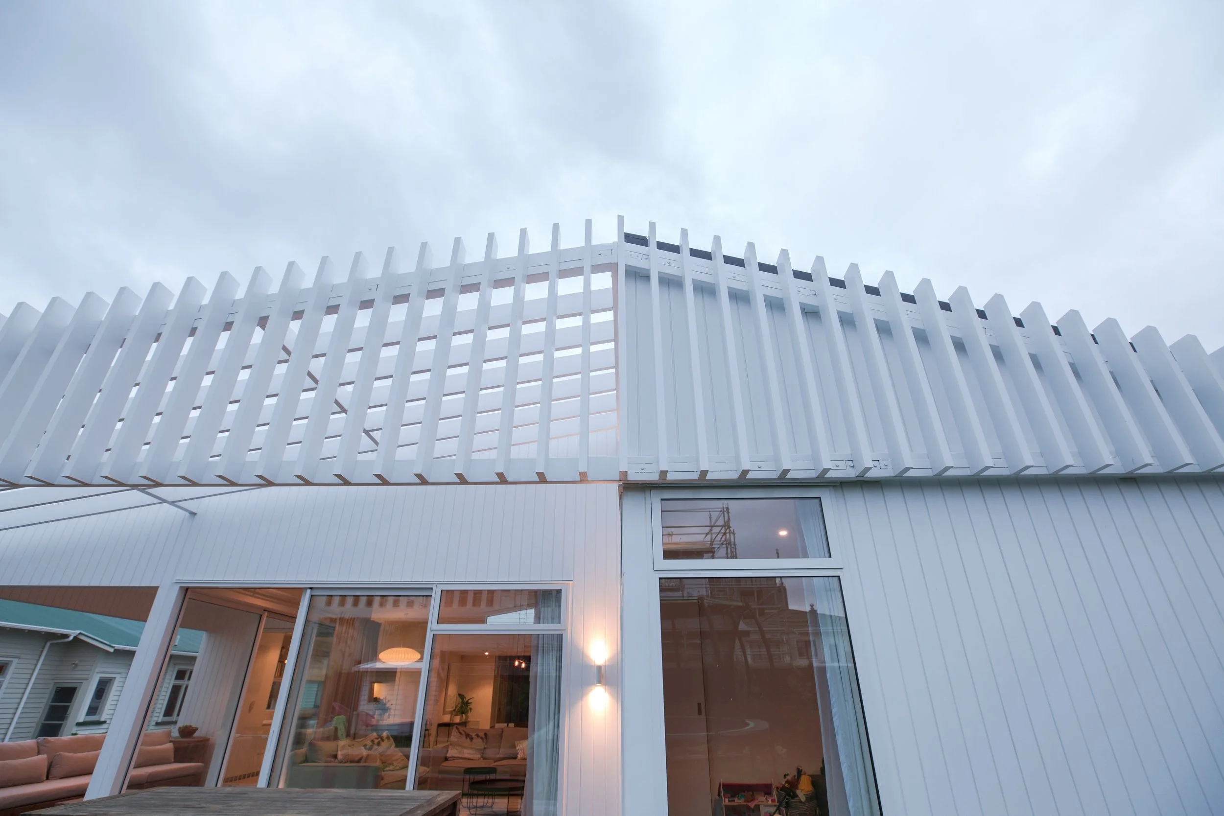 Modern white house with large windows and unique architectural design, featuring a distinctive slatted exterior structure on the upper level, against a cloudy sky background.