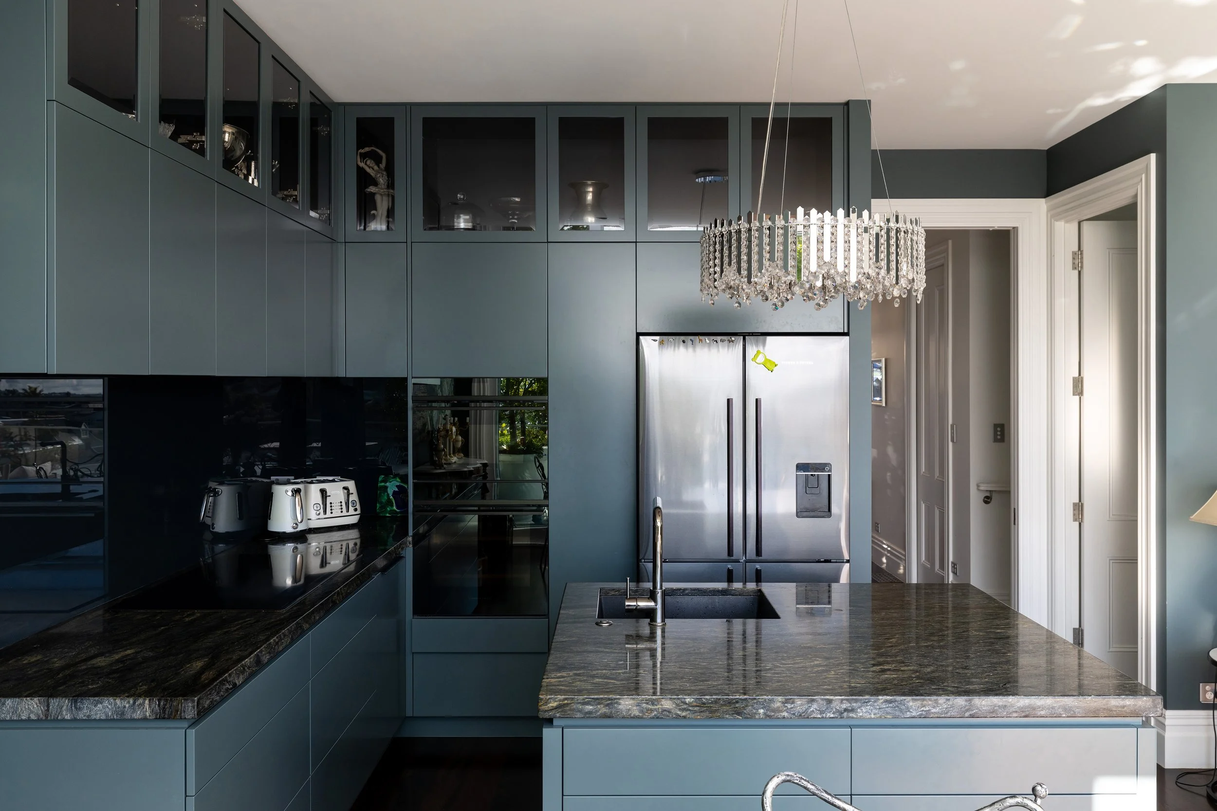 Modern kitchen with blue cabinets, granite countertops, stainless steel refrigerator, built-in oven, black backsplash, and a chandelier hanging above the island.