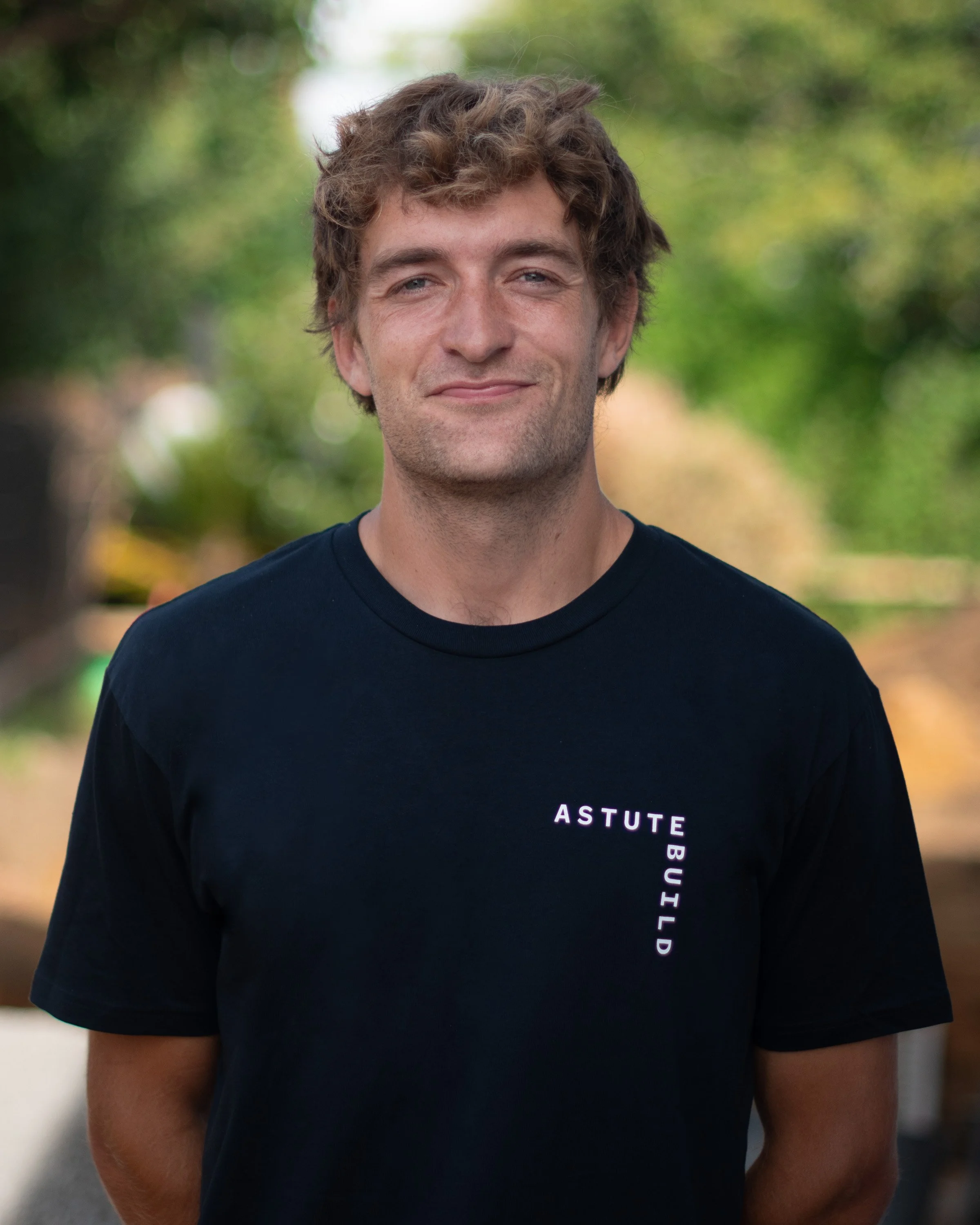 A young man with wavy brown hair and fair skin standing outdoors on a path with blurred trees in the background, wearing a black t-shirt with the text 'ASTUTE BUILD' printed on it.