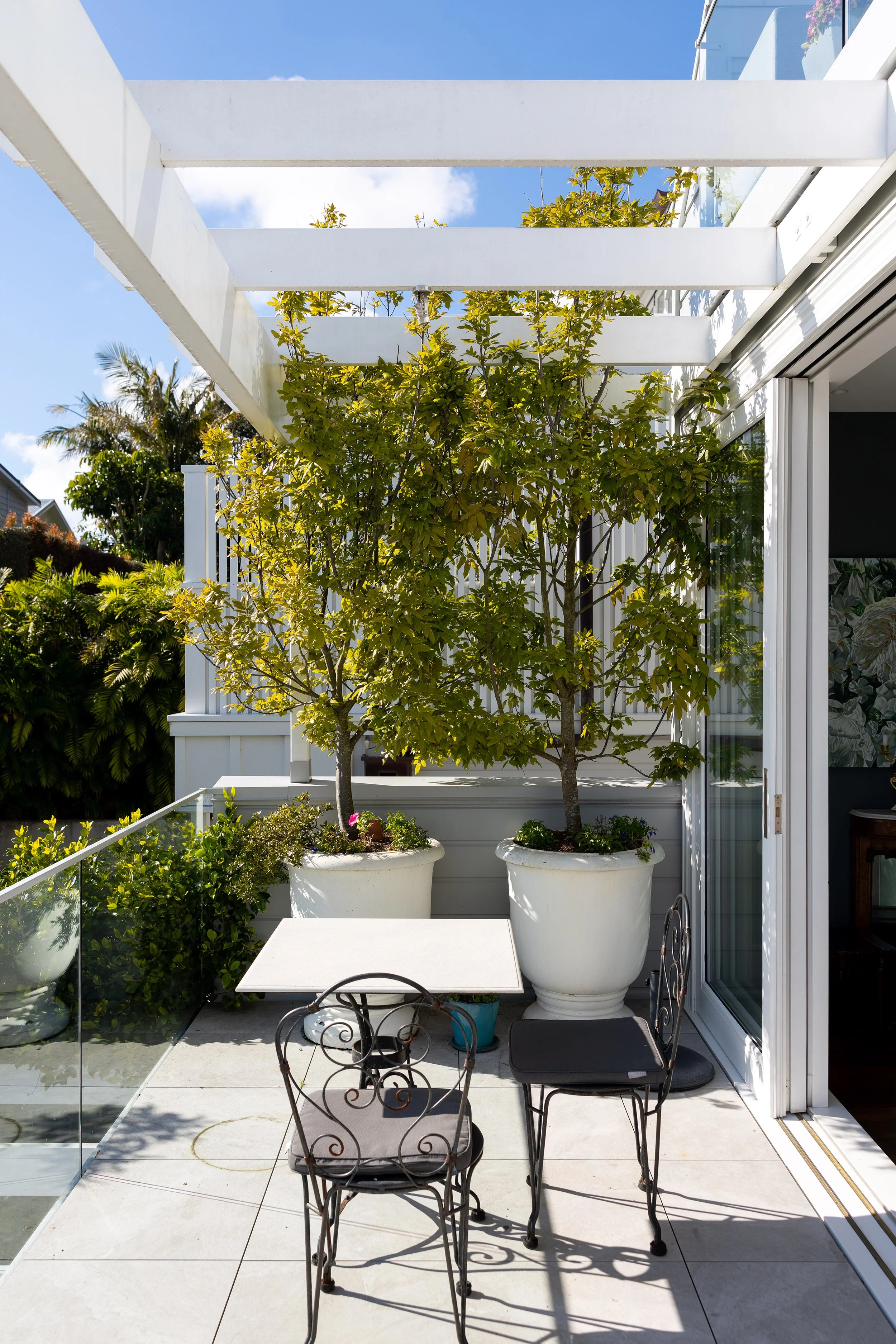 A small outdoor patio with two large potted trees, a white table, and two black wrought iron chairs. There are green shrubs and trees in the background, with a blue sky overhead.
