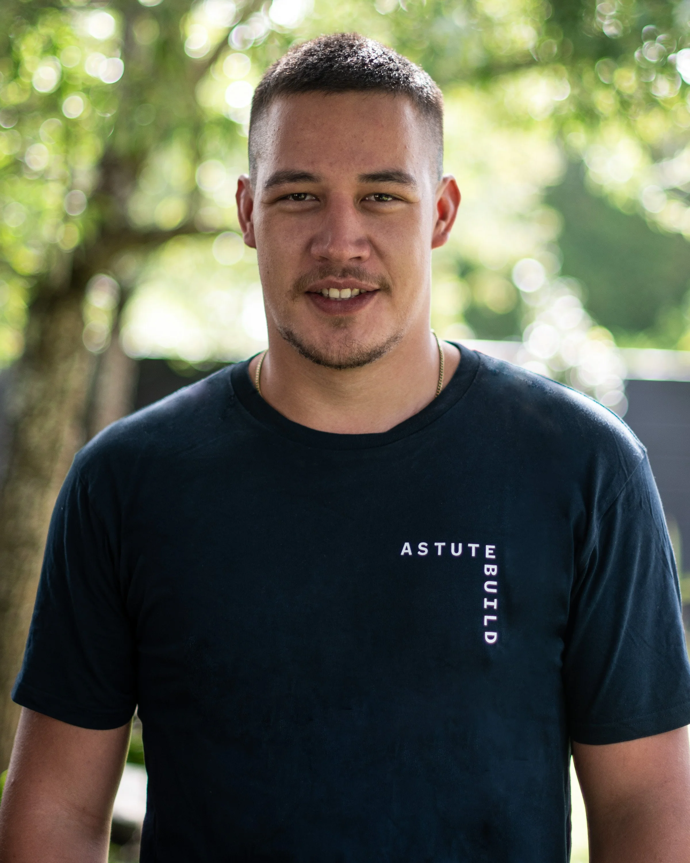 A young man with short dark hair, light facial hair, and a slight smile, wearing a black T-shirt with the words 'ASTUTE BUILD' printed vertically on the left side, standing outdoors with a blurred background of trees and greenery.