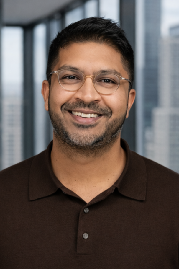 A smiling man with short dark hair, glasses, and a beard, wearing a dark polo shirt, standing in front of tall glass windows in a modern office building.