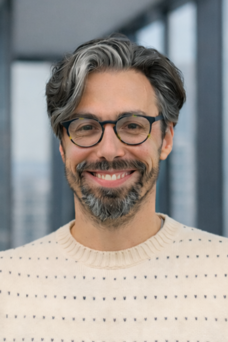 Smiling man with glasses and salt-and-pepper hair in an office with large windows.