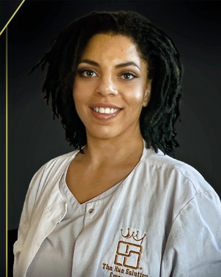 A young woman with shoulder-length curly black hair, wearing a white shirt with a logo that reads 'The Hua Solutions' and a crown symbol, smiling against a dark background.