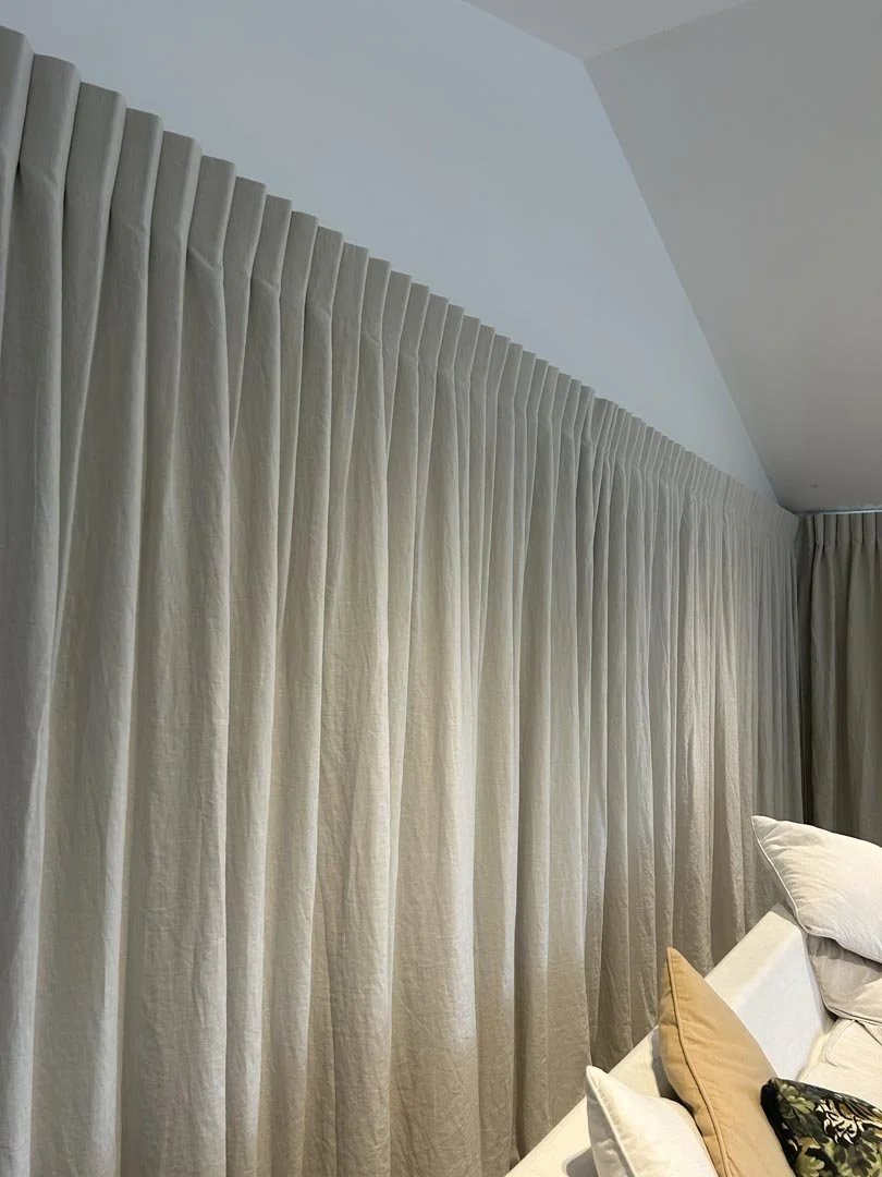 A set of beige curtains hanging over a window in a room, with a part of a bed and pillows visible in the lower right corner.