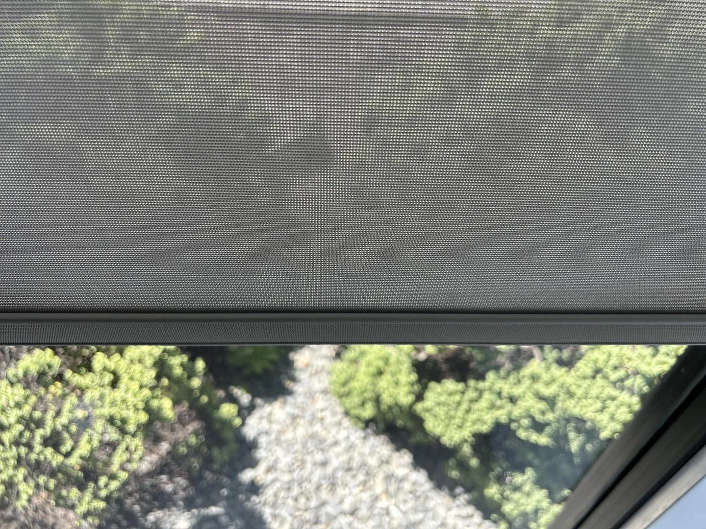 View through a window shows a blurry outdoor scene with green bushes and a gravel path, partially obscured by a grey window screen.