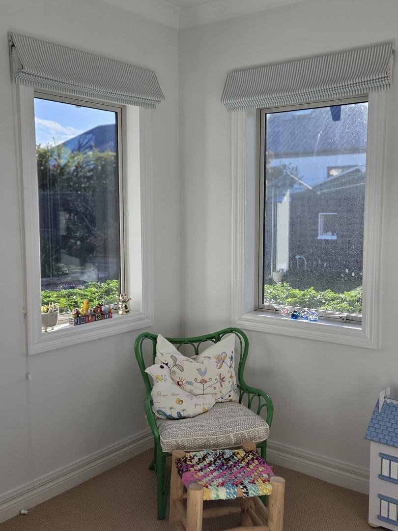A corner of a room with two windows, a green wooden chair with decorative pillows, a small wooden stool with a colorful woven cushion, and a windowsill with small decorative items.