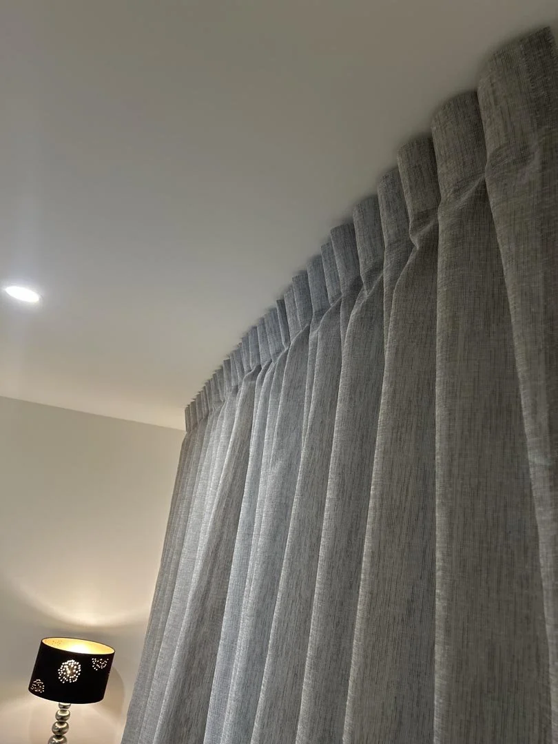 Gray curtains hanging from a window with a black table lamp with a floral cutout pattern on a side table beside it.