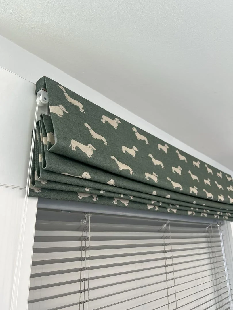 A green window valance with a pattern of beige dachshunds hangs above white horizontal blinds in a room.