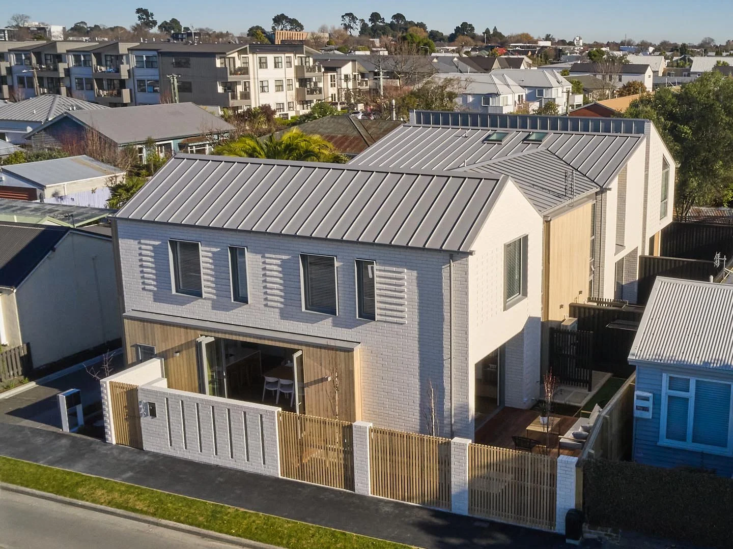 Modern two-story house with white brick and wood siding, metal roof, and fenced yard in a suburban neighborhood.