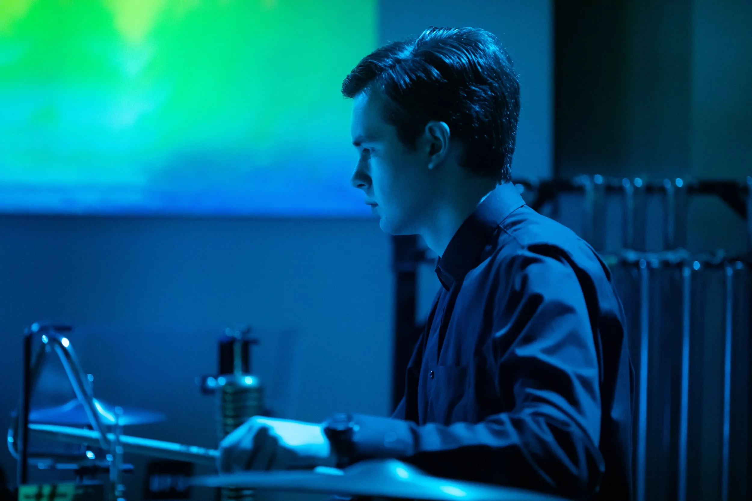 A young man with dark hair in a side profile working in a stage-lit percussion set up, with colorful digital displays in the background.