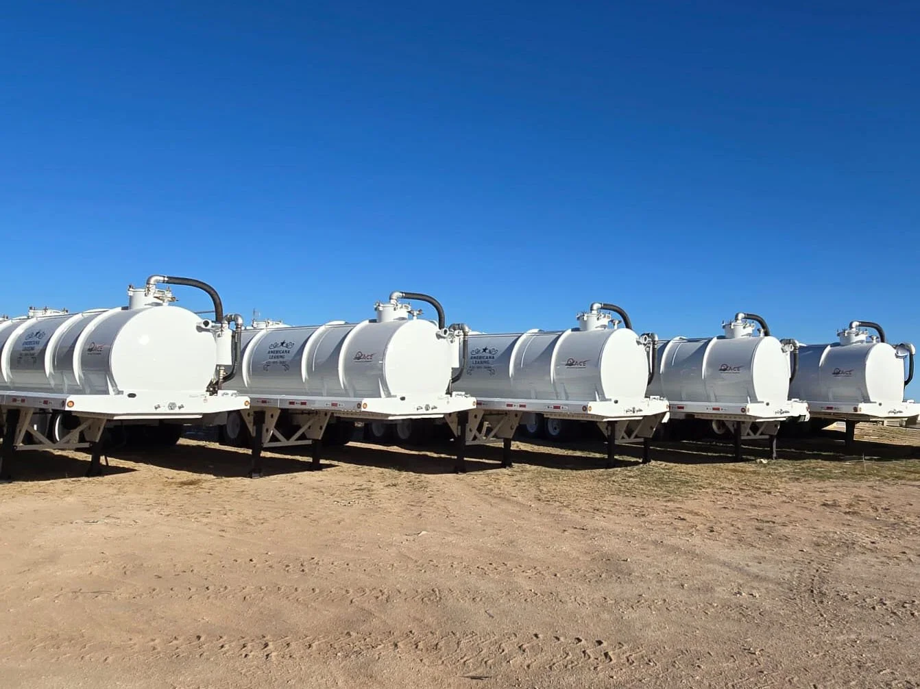 Six large white tanker trailers aligned on a dirt surface under a clear blue sky.