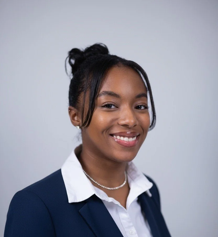 Portrait of a smiling young woman with dark hair styled in a bun, wearing a navy blazer, white shirt, pearl necklace, and earrings against a plain light gray background.