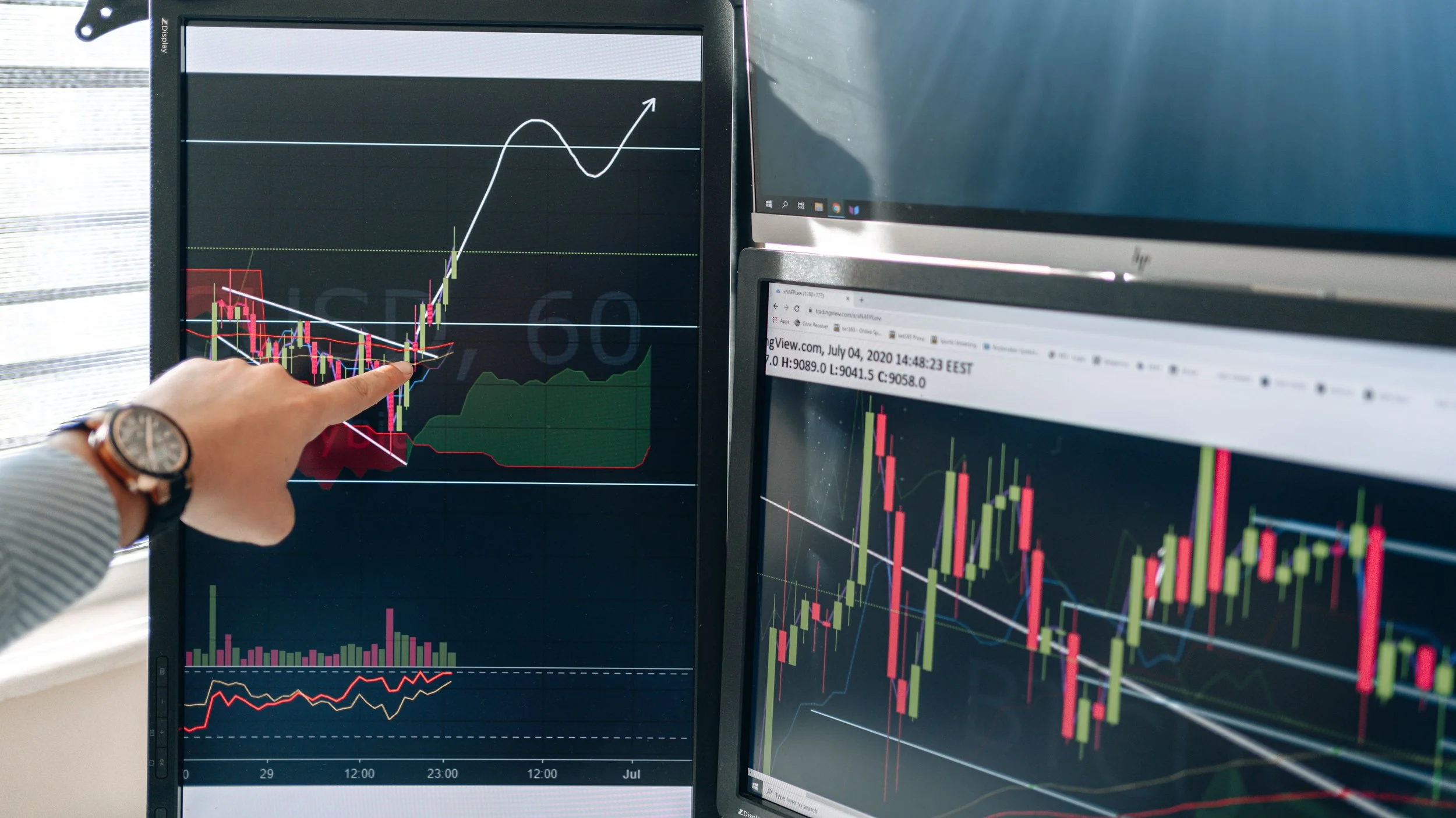 A person points at a stock trading chart with multiple other monitors displaying financial data in the background.