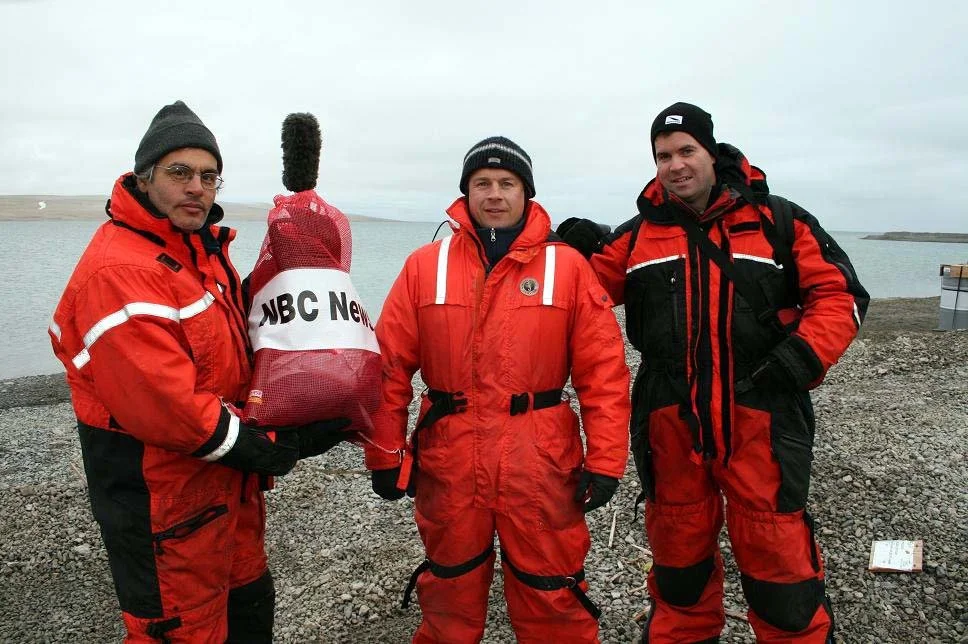 Three men in red and black outdoor jackets standing on rocky shoreline near water, holding a red NBC News bag, with an overcast sky and distant land in the background.