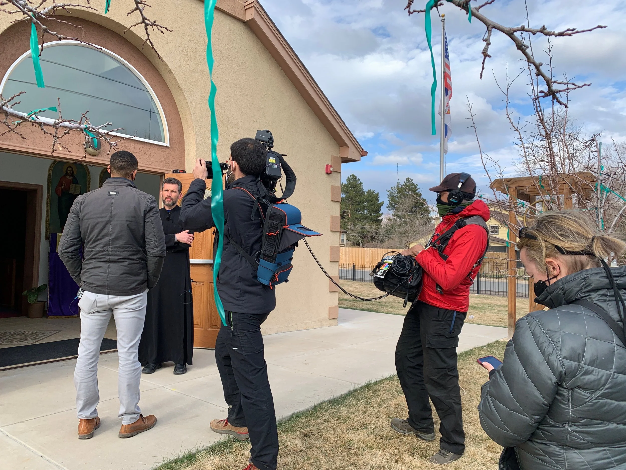 A group of people including a clergyman, journalists, and videographers gathered outside a church or religious building, with some recording and taking pictures during a ceremony or event. The building has an arched window and a flag flying outside.