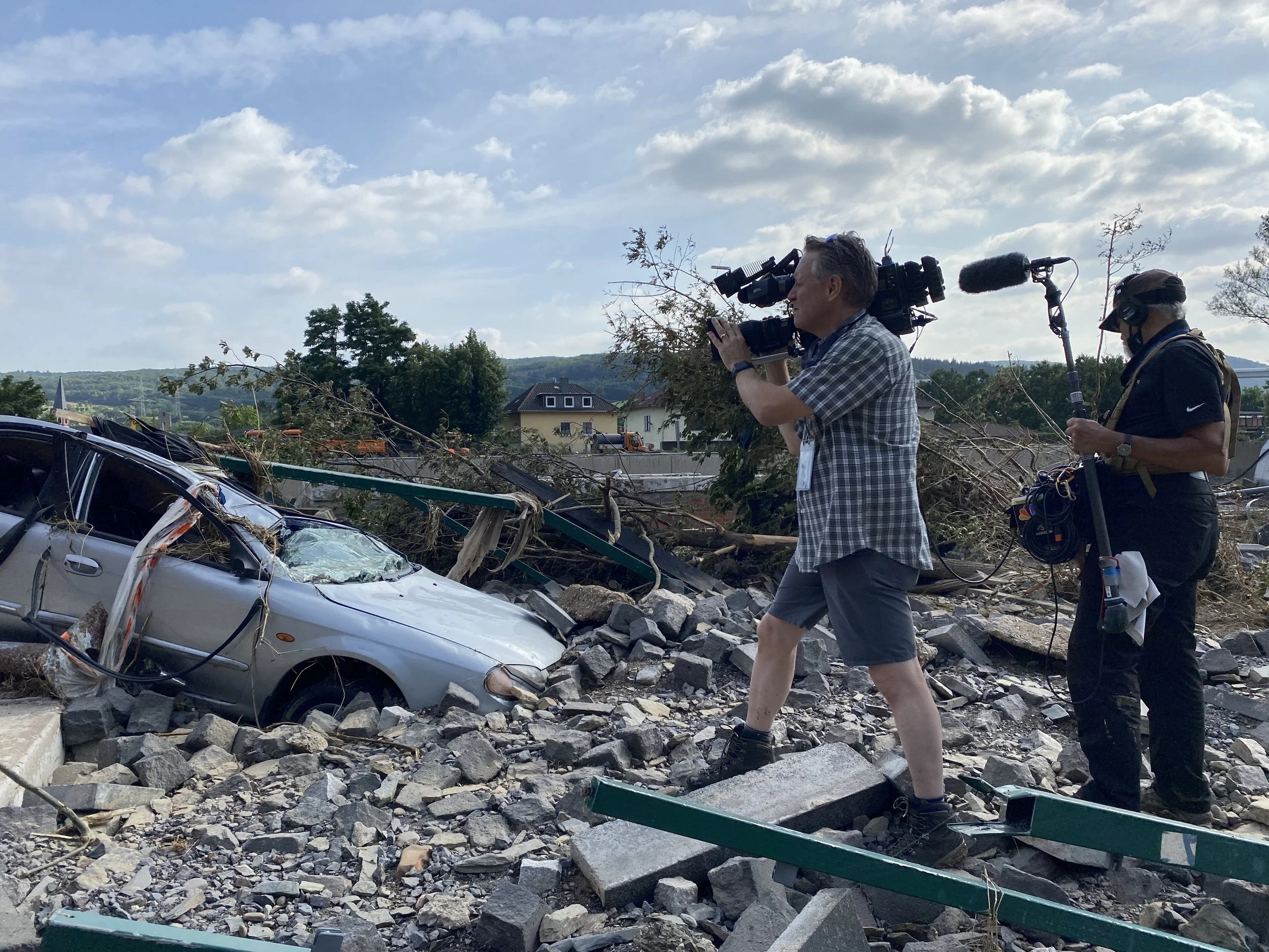 A news crew filming a scene of a car accident with debris and fallen trees after a storm or disaster, on a partly cloudy day.